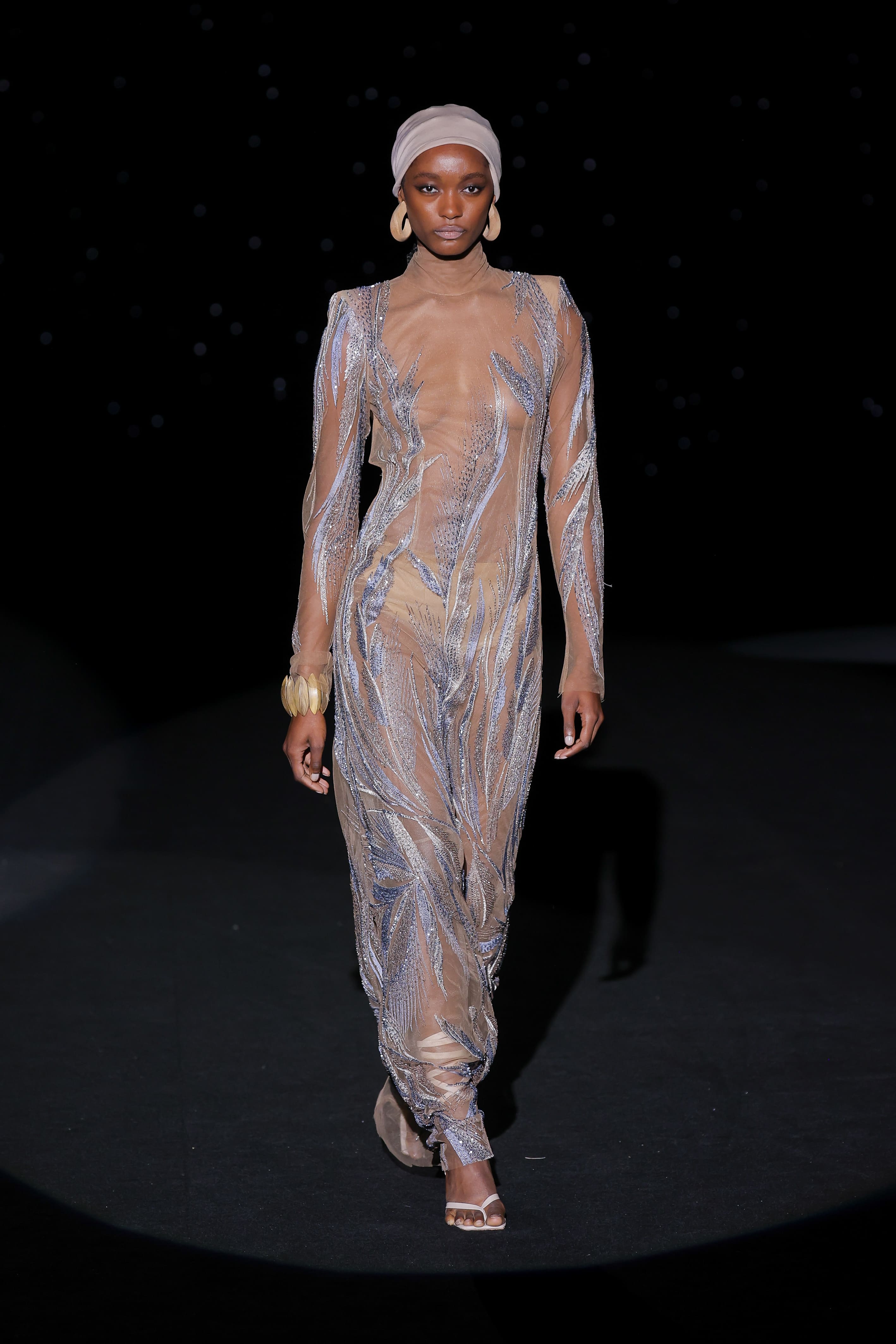 look is an ode to the body as a canvas and the dress as a trace of light. The design, built on a transparent nude base, adheres to the silhouette with the delicacy of a sigh, while embroidered sequins in shades of blue and silver draw organic shapes reminiscent of spikes or branches in the wind.