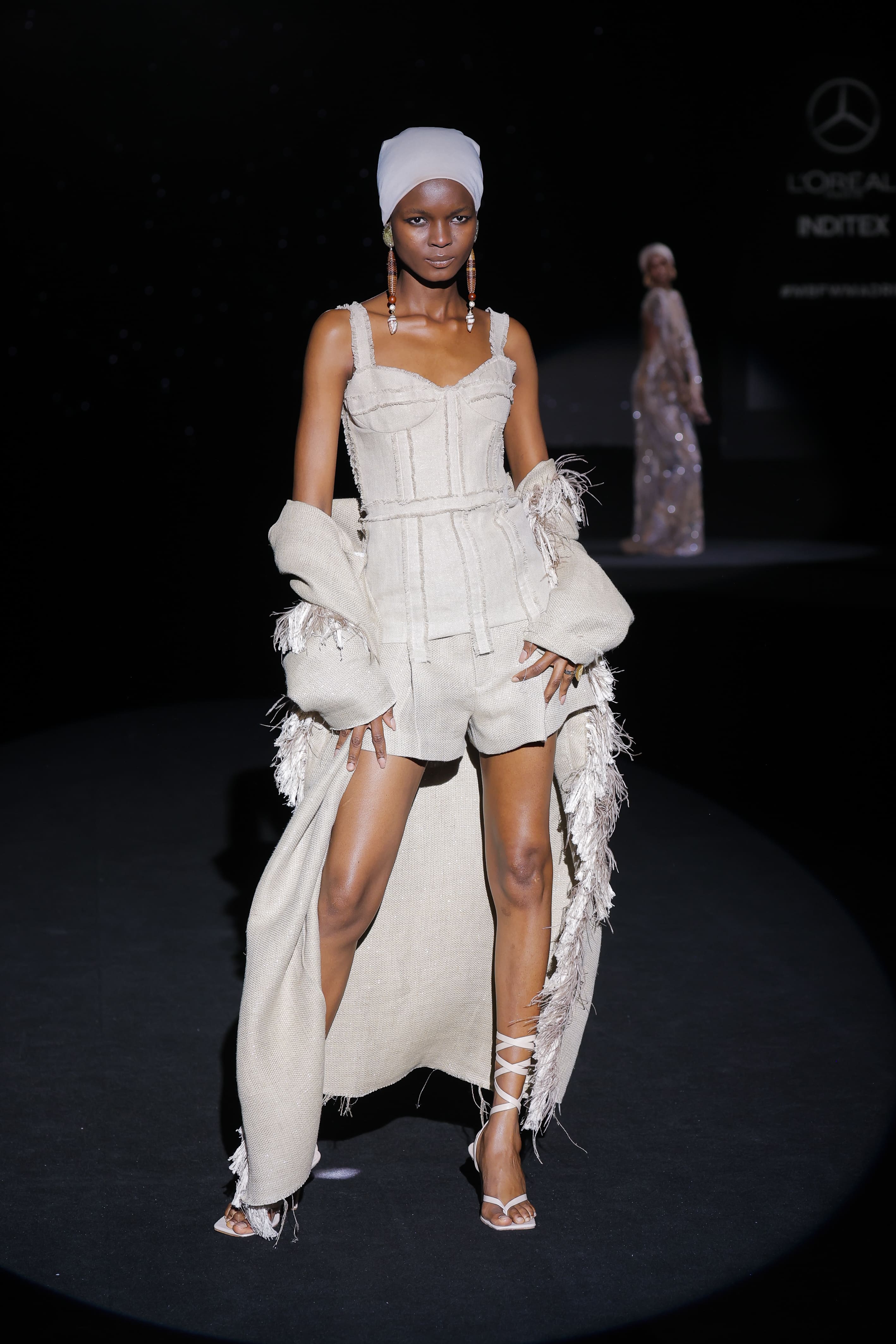image confirms the ritual and earthy essence of the look: the body becomes a textile sculpture. The close-up reveals the details of the craftsmanship: the structured corset with external stitching, the raw texture of the fabric and the fringes that run through the cape like roots in the wind.