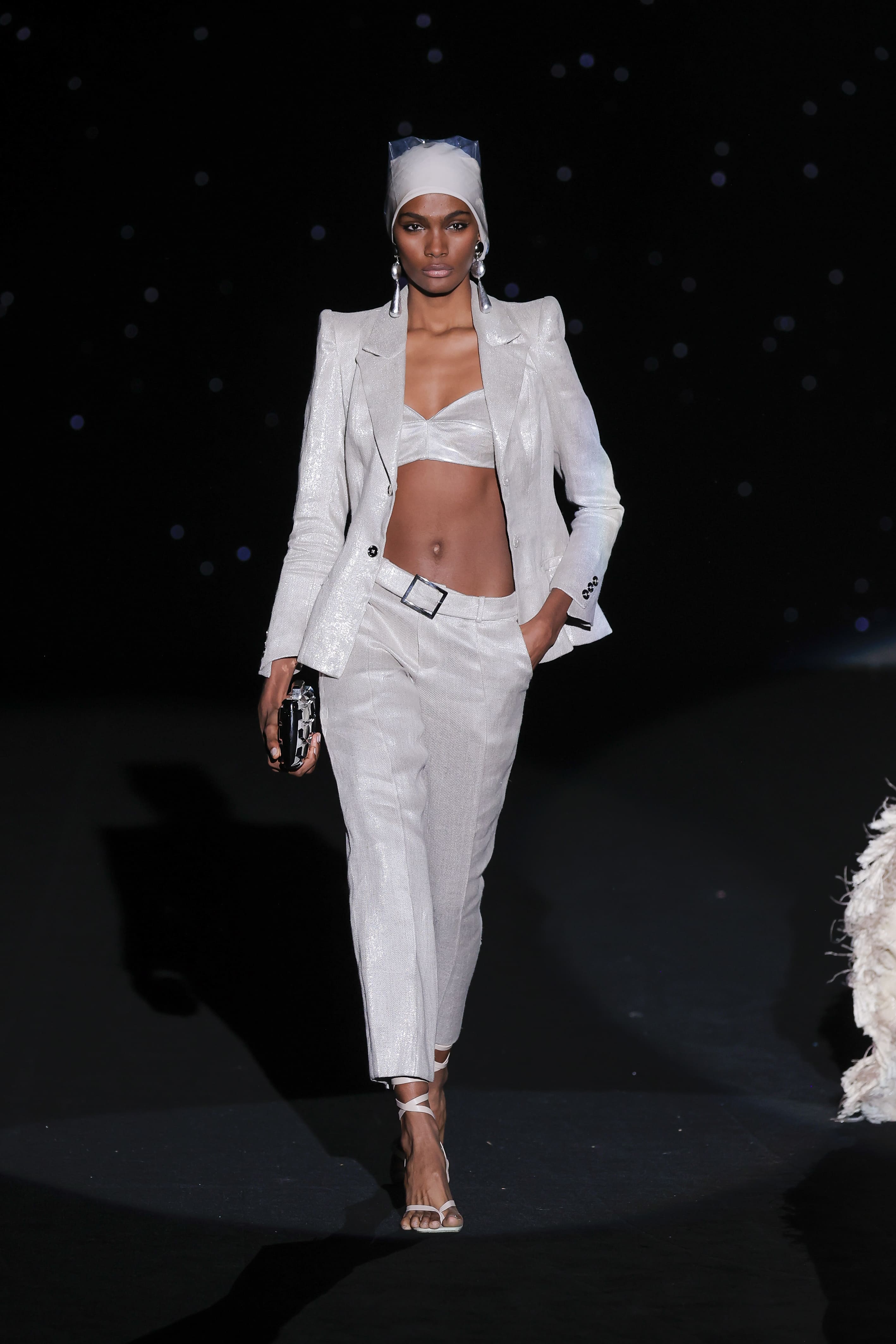 look reinterprets the classic suit jacket with a luminous and feminine power. The pearly white ensemble, impeccably cut and woven with a subtle metallic sheen, is elevated to the max by the cropped bustier, which breaks up the formality with precise sensuality. The high waist and belt mark the silhouette with architectural elegance.