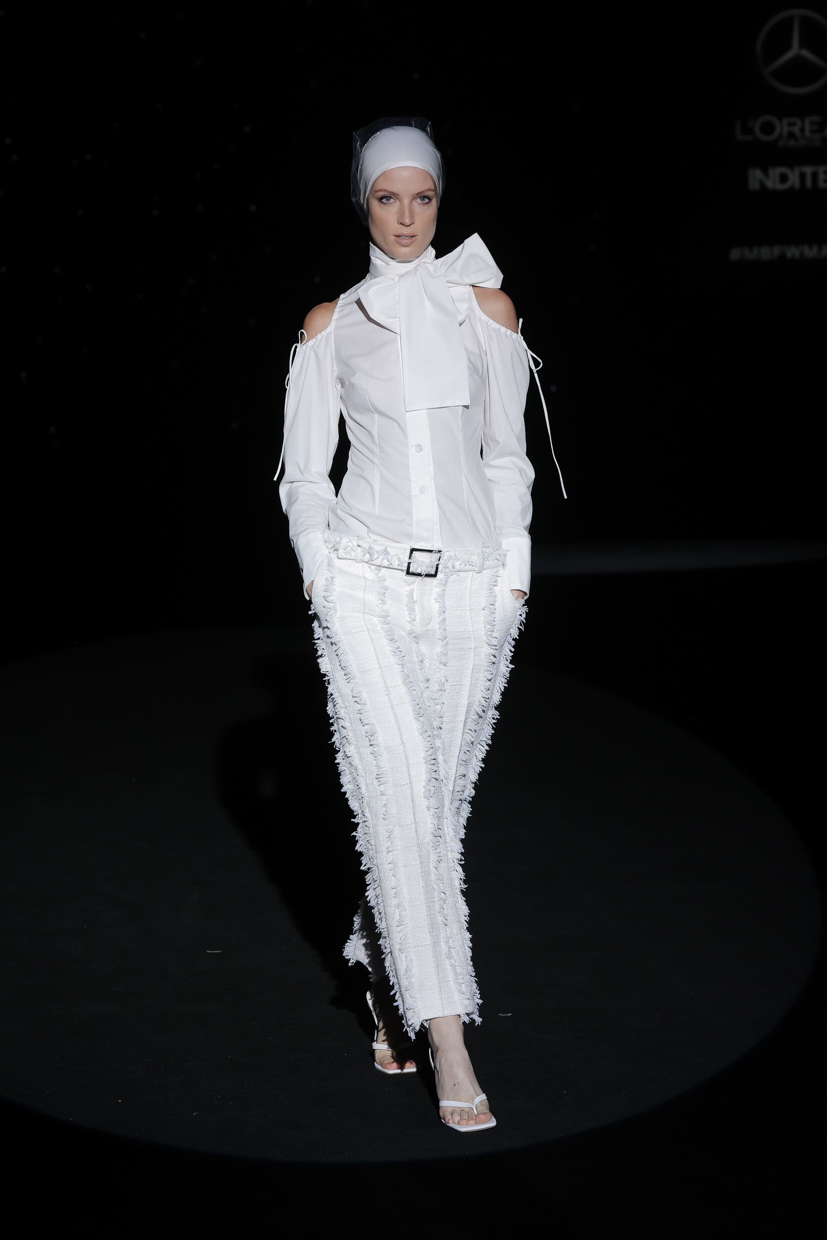 This white ensemble reinterprets classicism with a contemporary twist, full of theatricality and subtlety. The structured shirt with bare shoulders and large bow at the neck evokes the sophistication of period costume, transformed here into a modern gesture of power.