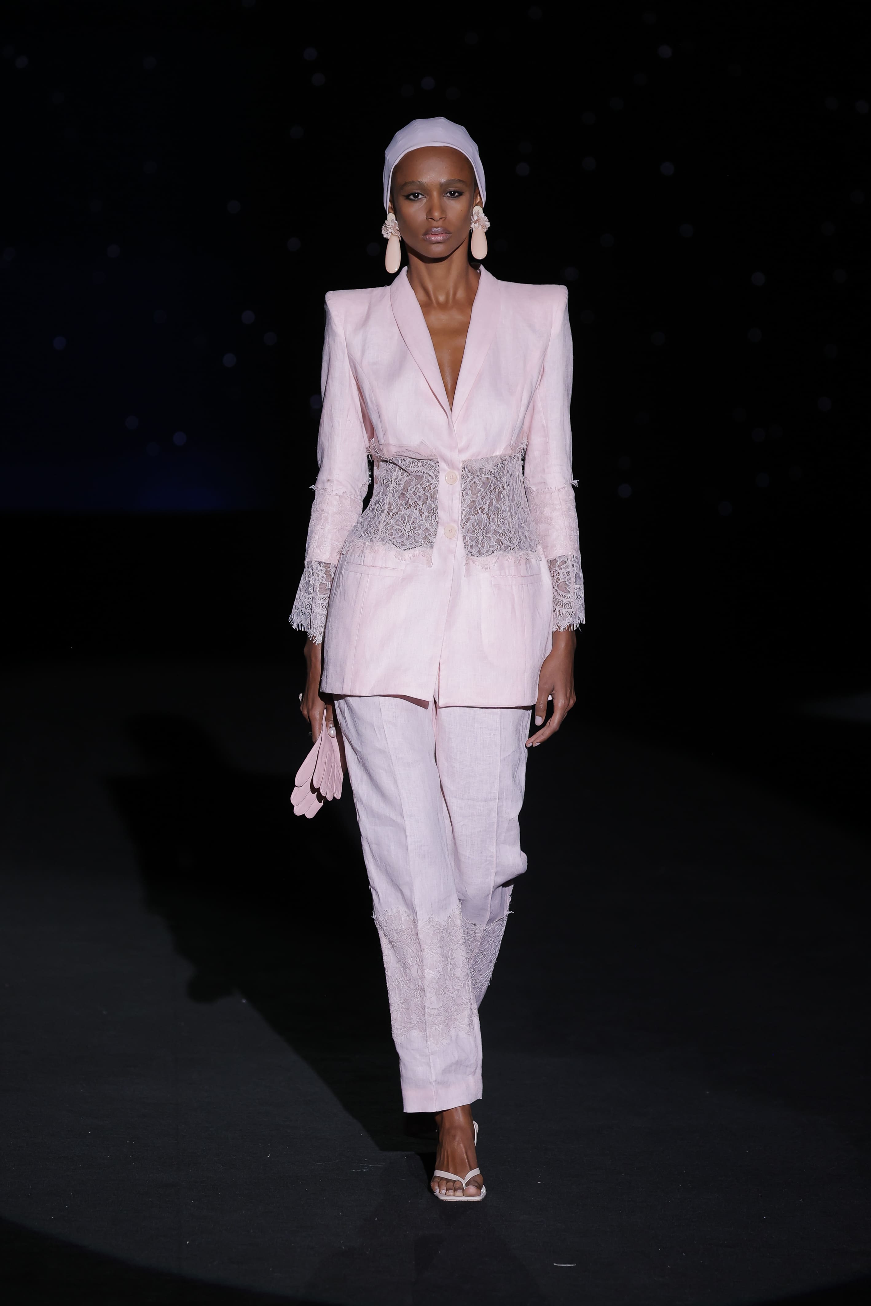 This powder pink ensemble is an ode to architectural femininity. The linen suit, with its impeccable structure and pronounced shoulders, contrasts with the delicacy of the lace that insinuates itself at the waist and subtly extends to the cuffs and bottom of the trousers, creating a sensual silhouette without sacrificing the solidity of the cut.