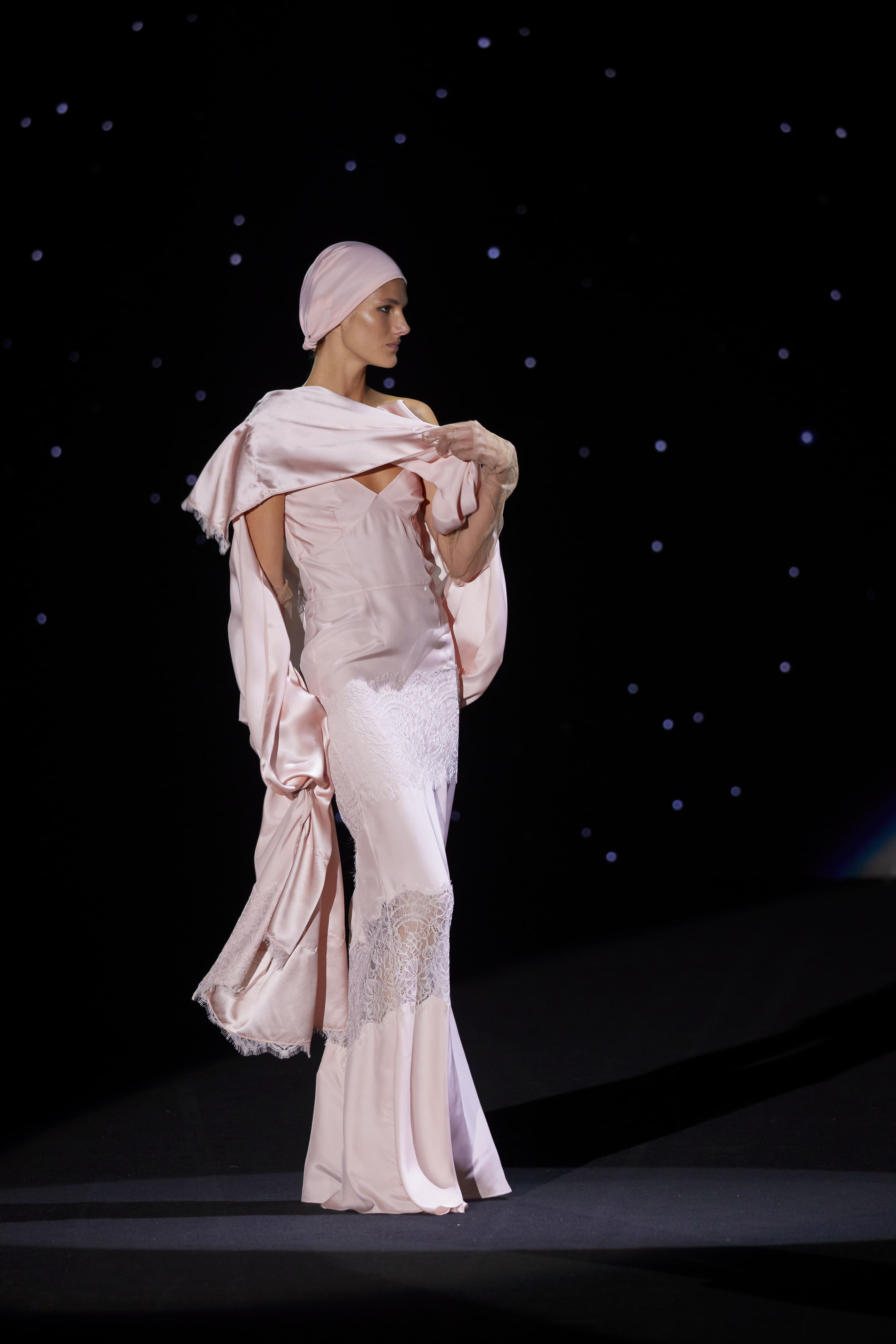 This long dress in powder pink satin dazzles with its classic, refined elegance. Its fitted cut accentuates the figure, while the delicate sheer lace panels on the skirt lend an ethereal and sophisticated air, creating a subtle contrast between opacity and transparency.