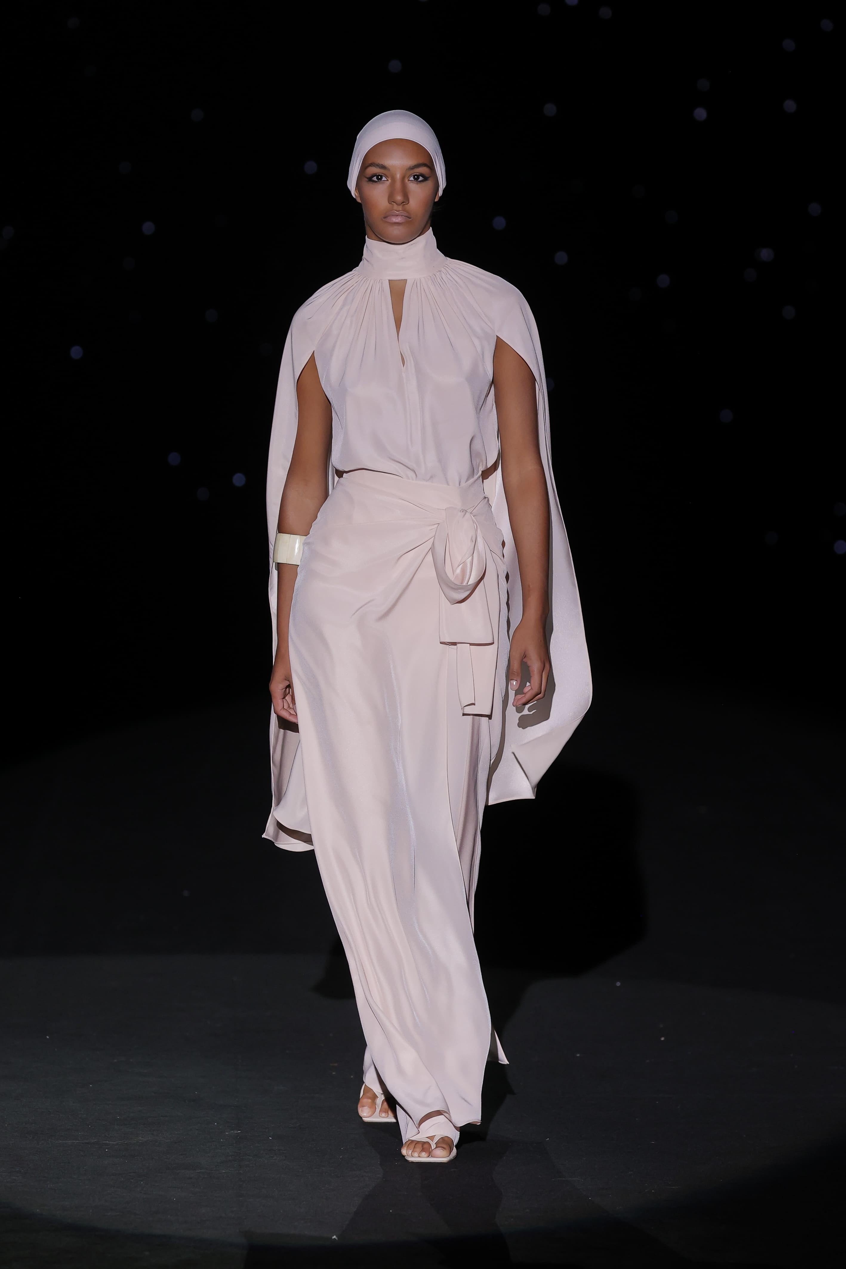 This elegant ensemble in soft pastel pink is characterised by its fluidity and sophistication. Crafted in a lightweight satin fabric, the blouse features a high neckline and delicate pleats for texture and movement, with a cape that falls gracefully over the shoulders, adding a dramatic and ethereal air.
