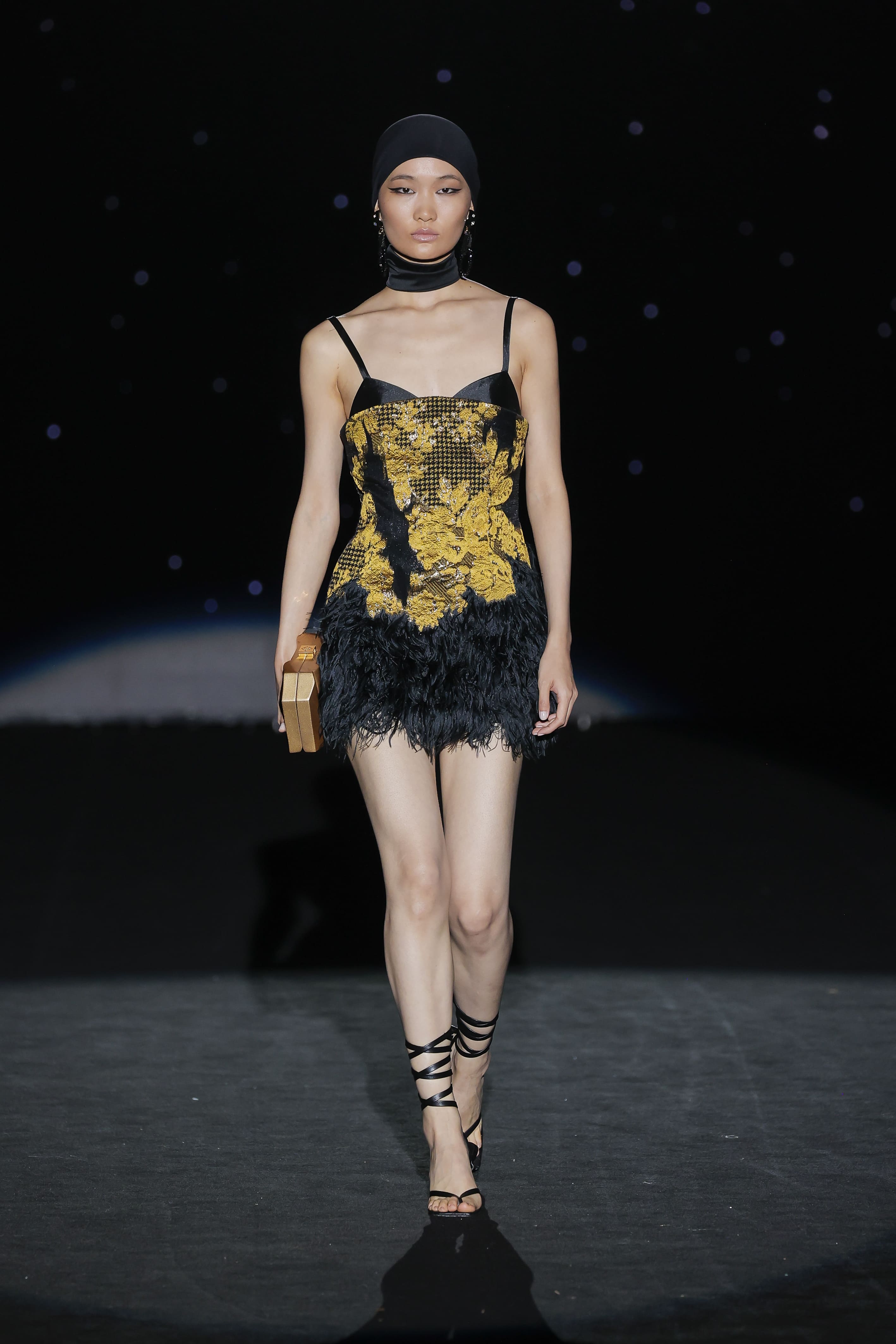 a breath of baroque theatricality and contemporary synthesis. This mini-dress with a lingerie neckline plays with the codes of excess and restraint: satin black becomes the backdrop for a profusion of golden embroidery evoking antique tapestries and imperial gardens, contrasting with the sensual lightness of the feather-covered hemline.
