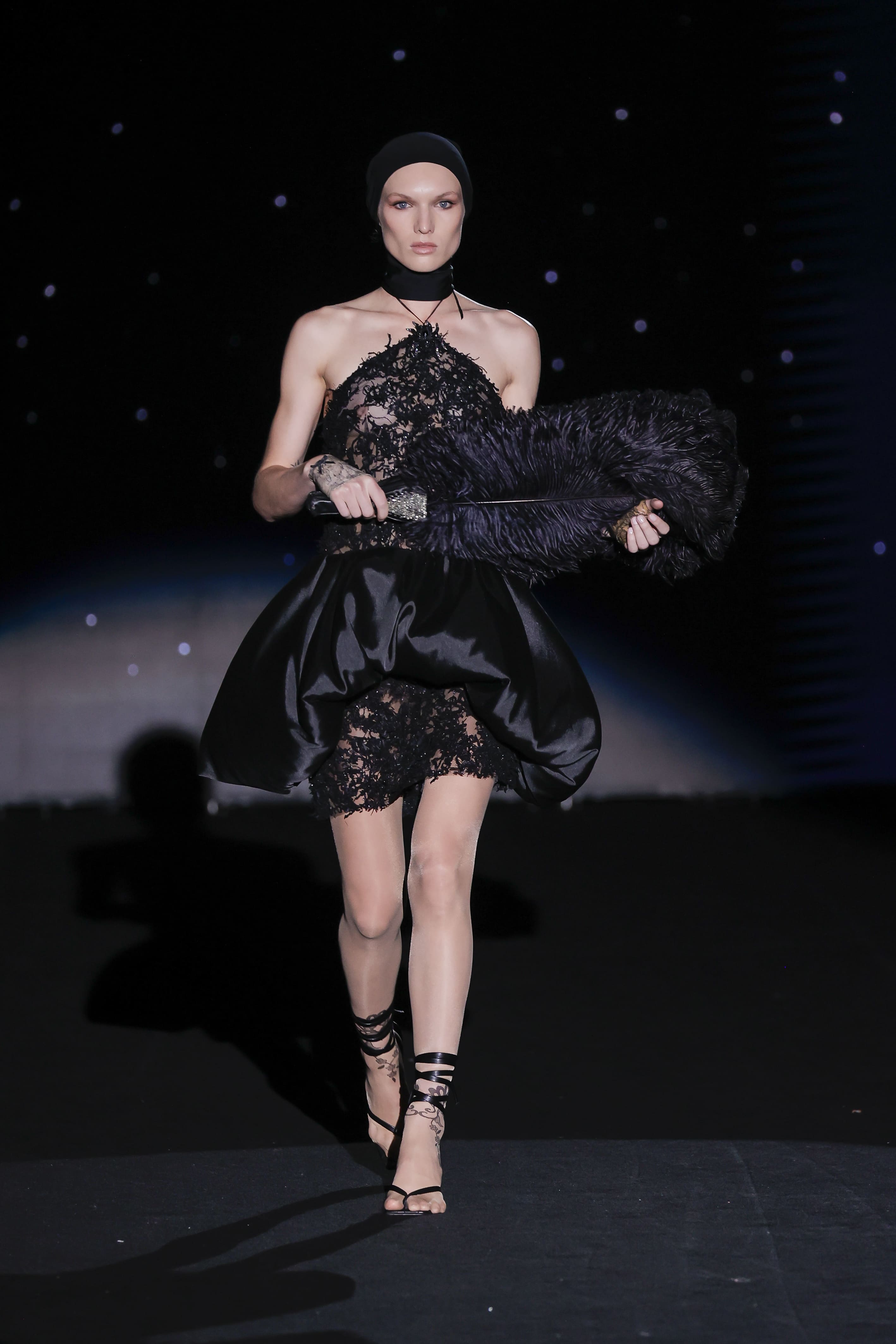 design is a burst of sophisticated theatricality and subversive femininity. The body is enveloped in organic, almost vegetal black lace that ascends to the neck like a wild vine. The halter bustier leaves the shoulders bare and frames the silhouette with sculptural precision.