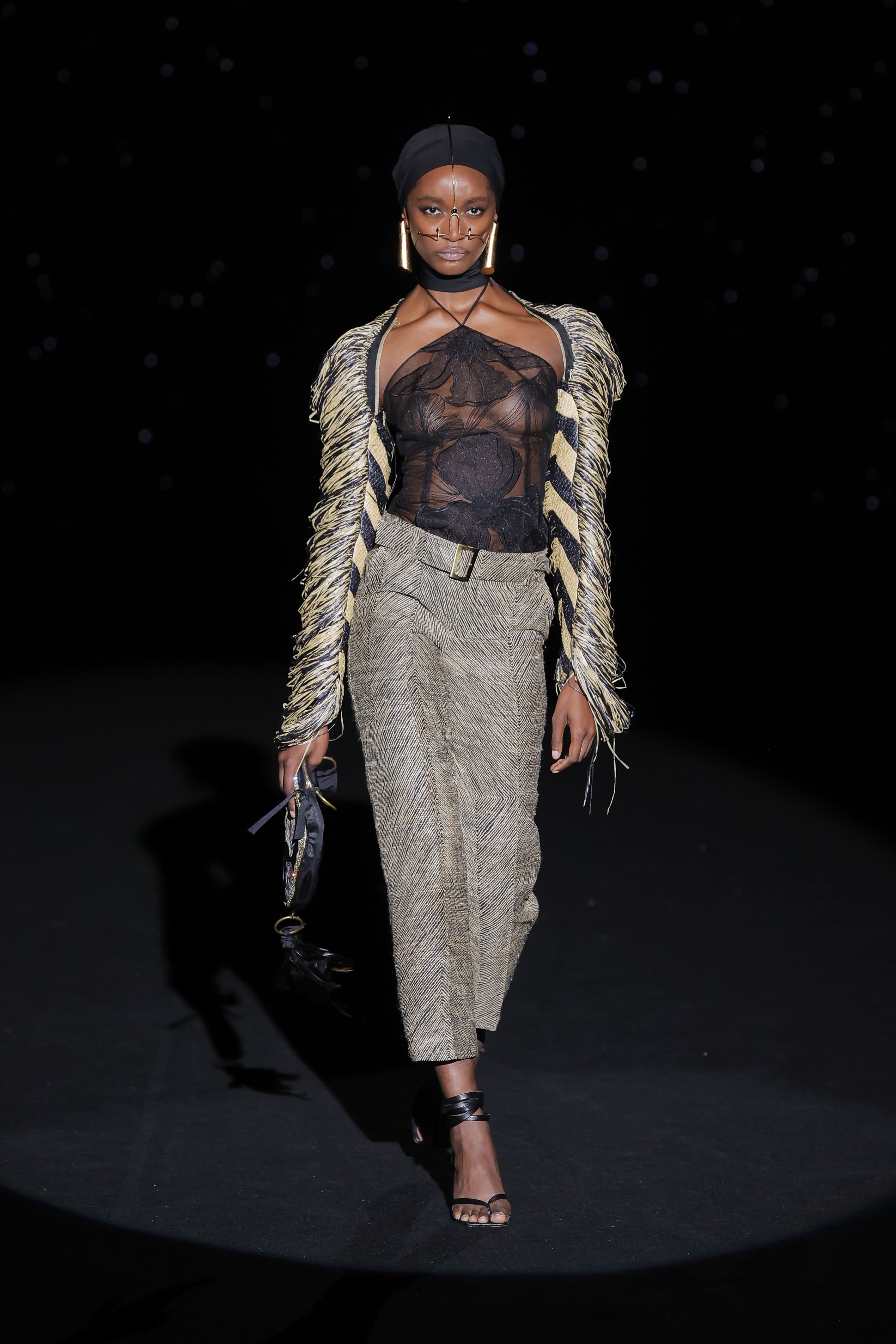 A magnetic figure that combines tribal strength and urban sophistication. The sheer top, embroidered with dark flowers, wraps around the torso like a veil that reveals more than it hides, while the black and gold fringed jacket falls like hand-woven armour.