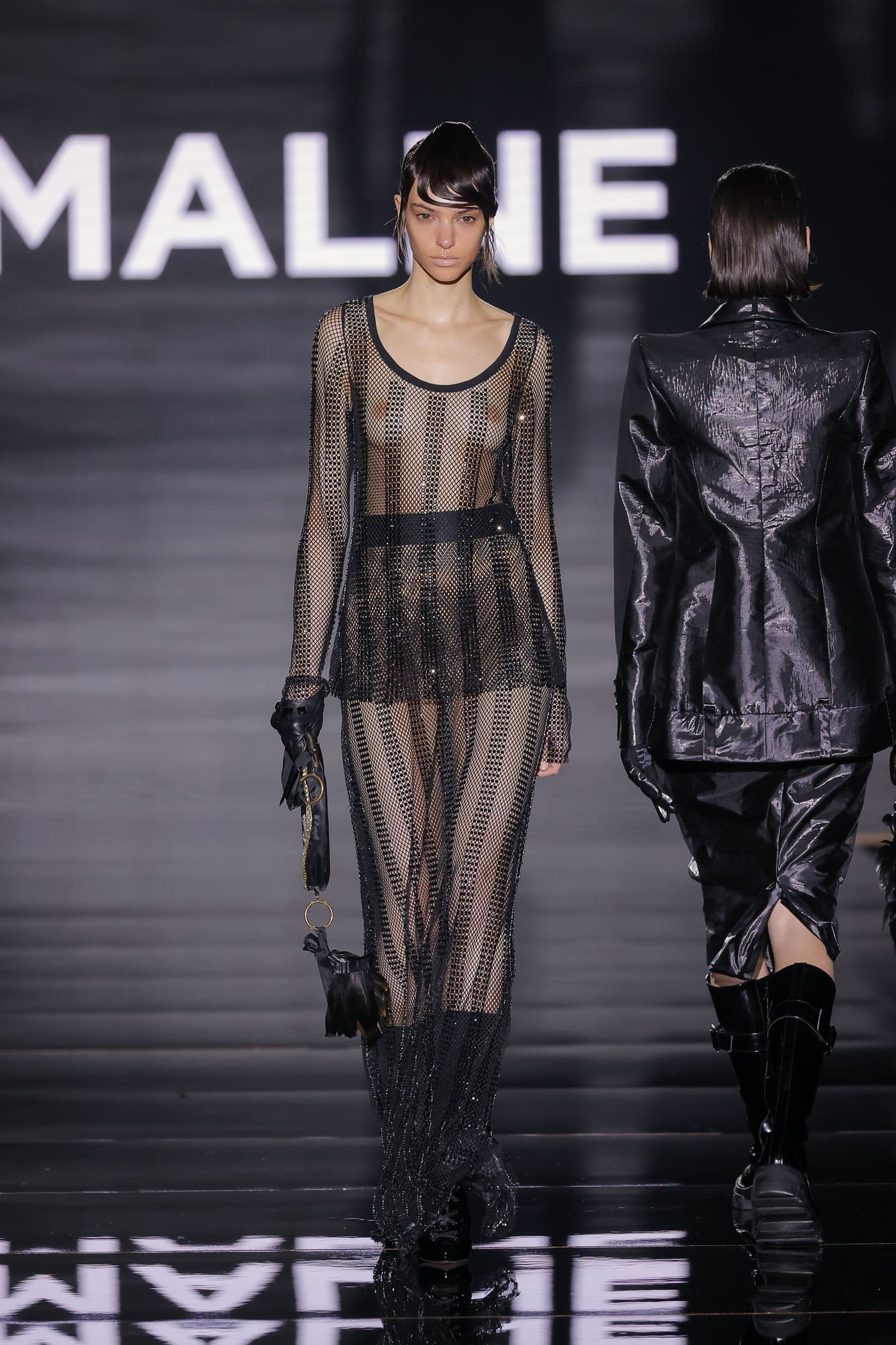 Model on the catwalk wearing a semi-transparent black dress by Malne covered in glitter, with long sleeves, a round neckline and a straight silhouette