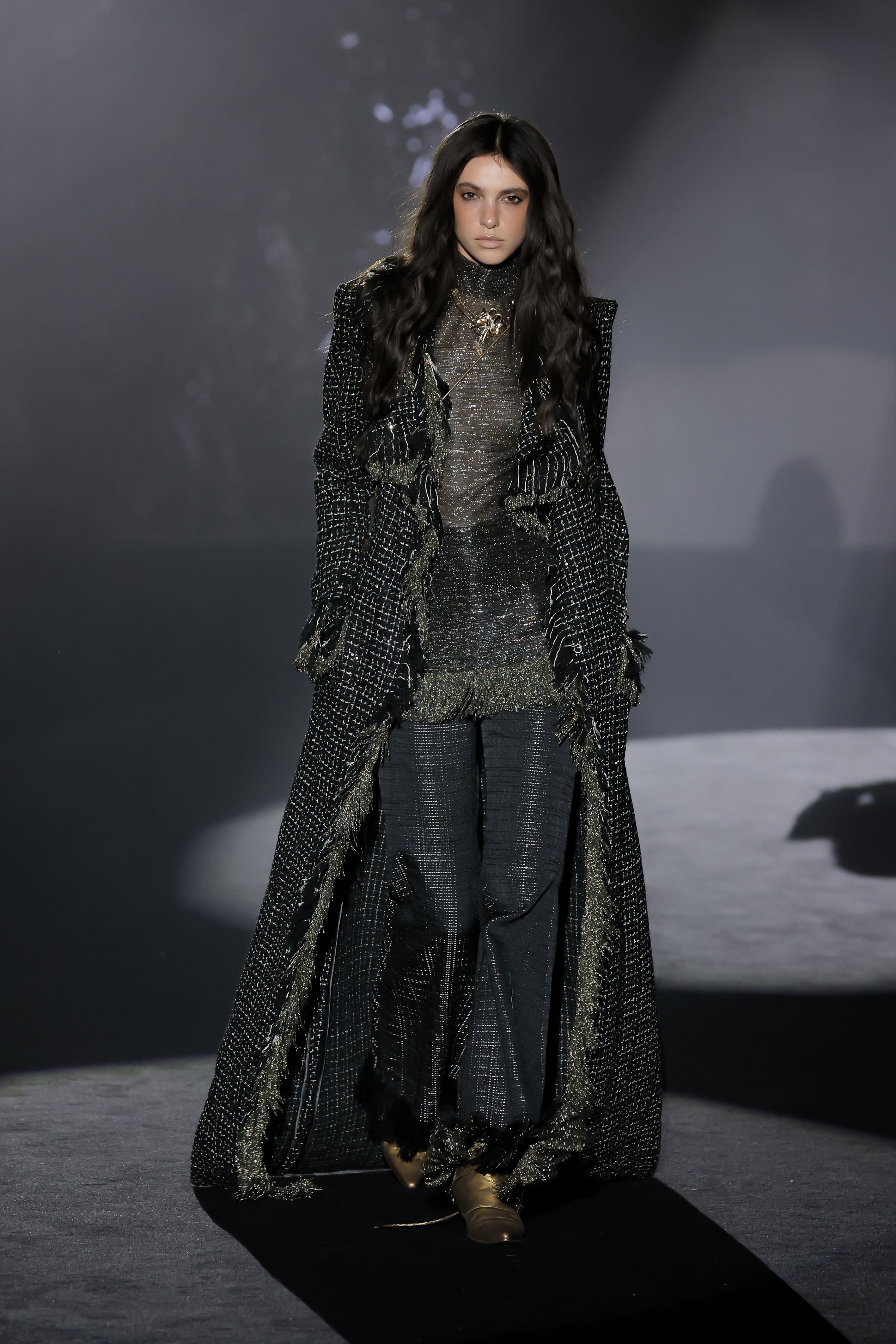Model walks the runway in a dark textured ensemble and long coat during a MALNE fashion show.
