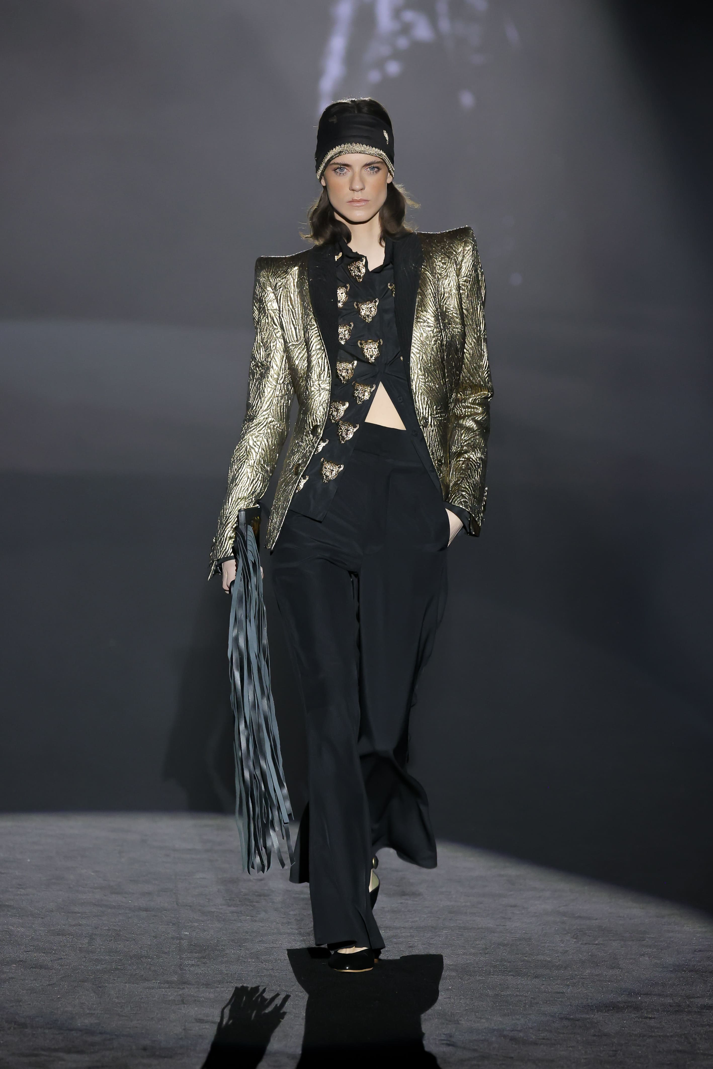 Model walks the runway in a shiny gold jacket and black trousers.