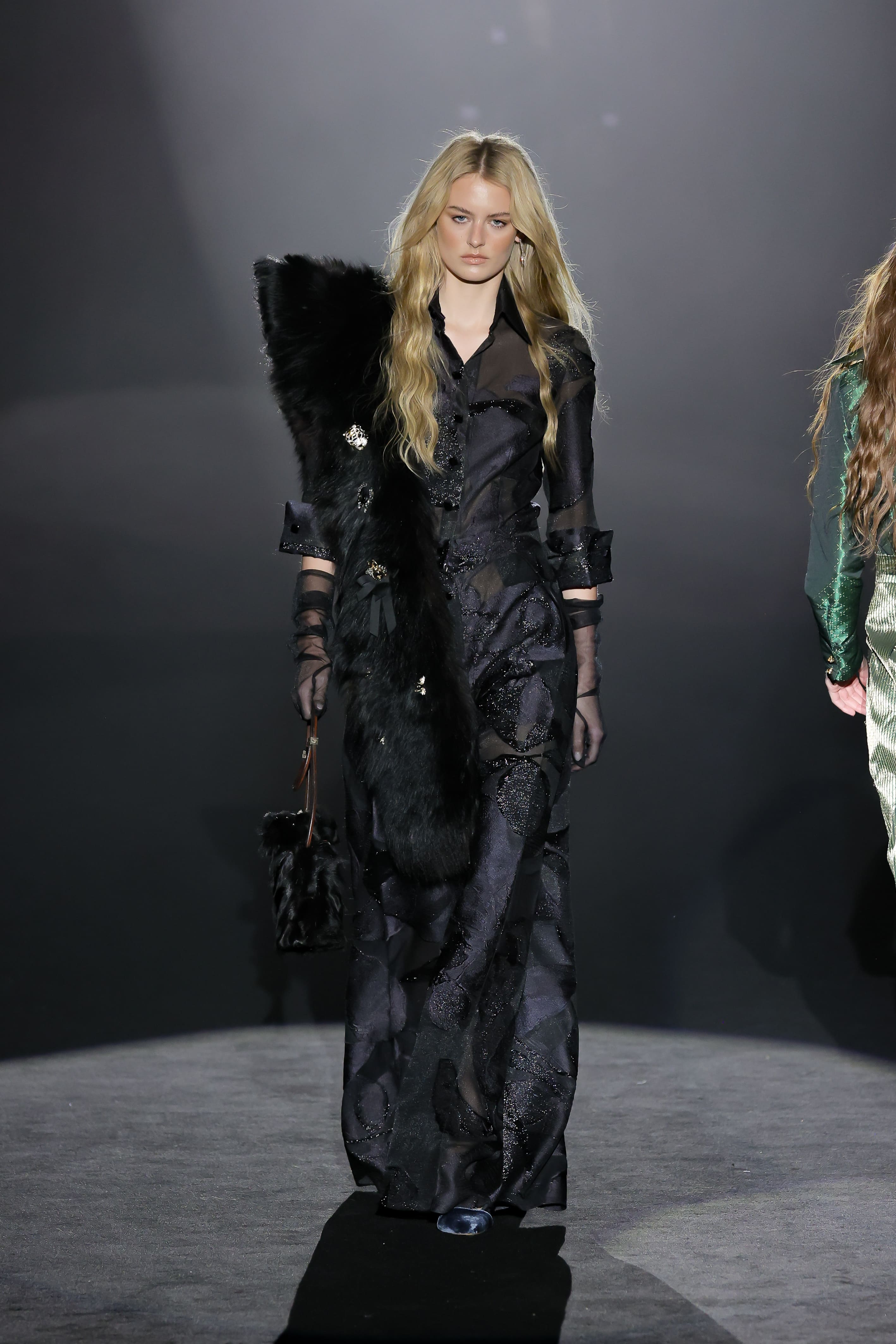 Model walks the runway in a long black dress with sheer panels and feather detailing