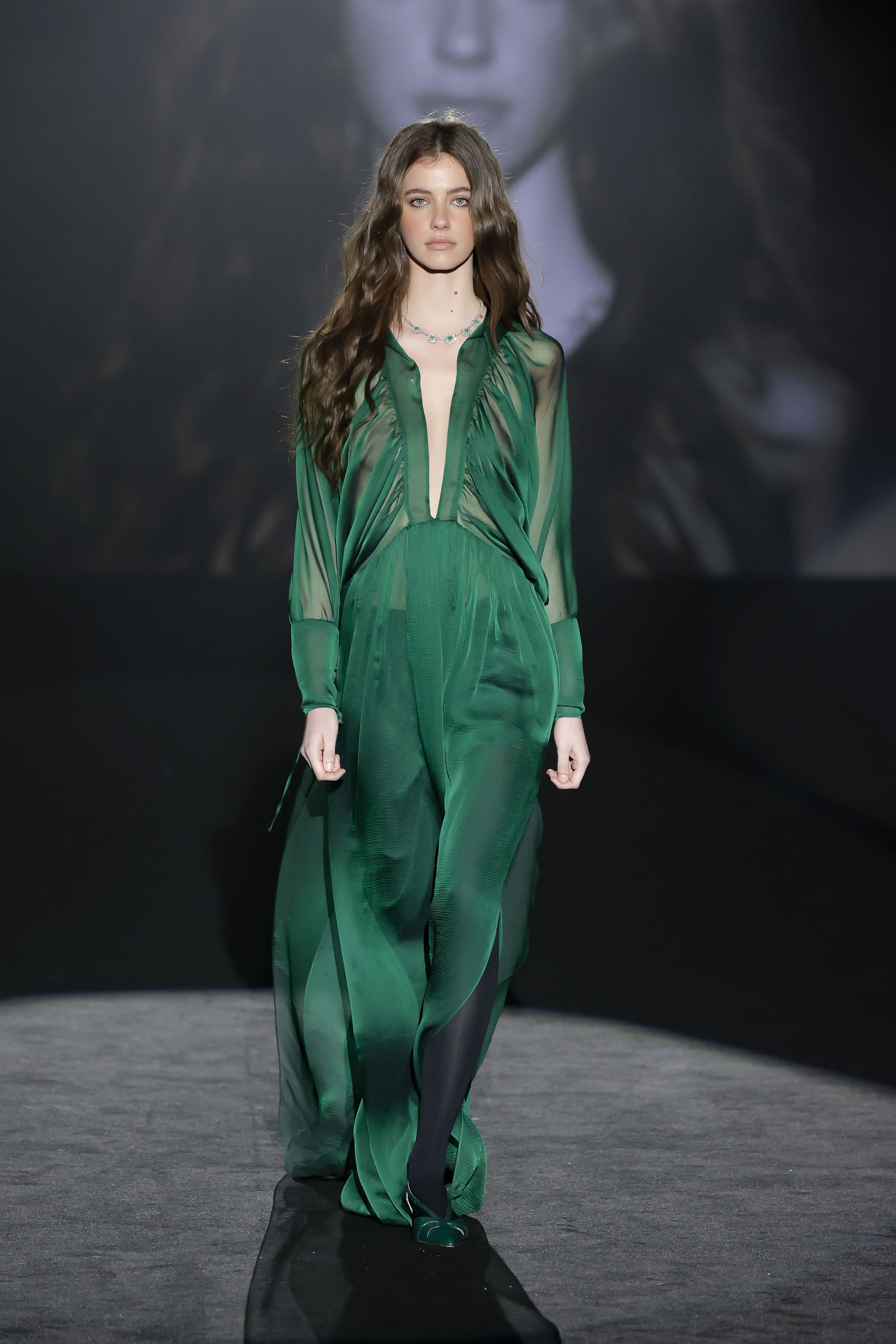 The model struts down the runway in a long, flowing green dress with a plunging neckline.