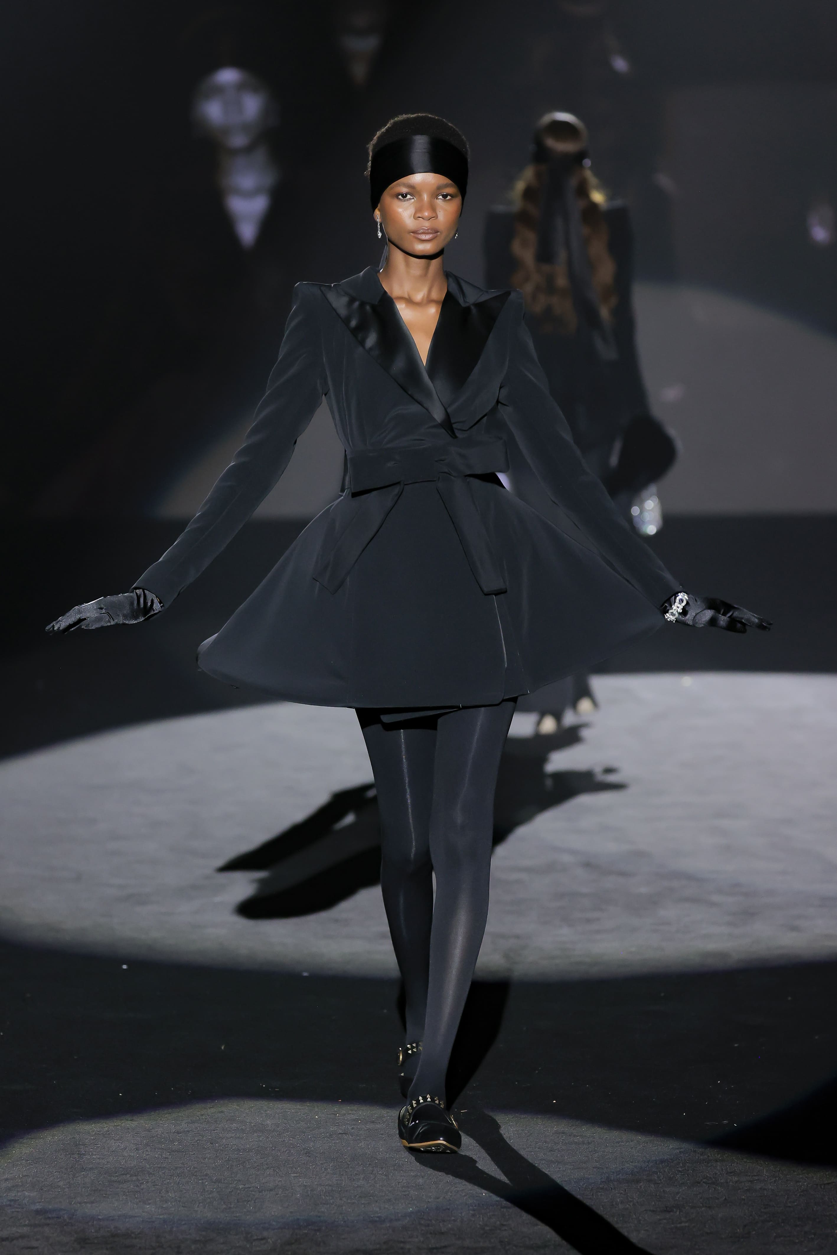 Model struts down the runway in a structured black outfit with a short skirt, gloves and a headband