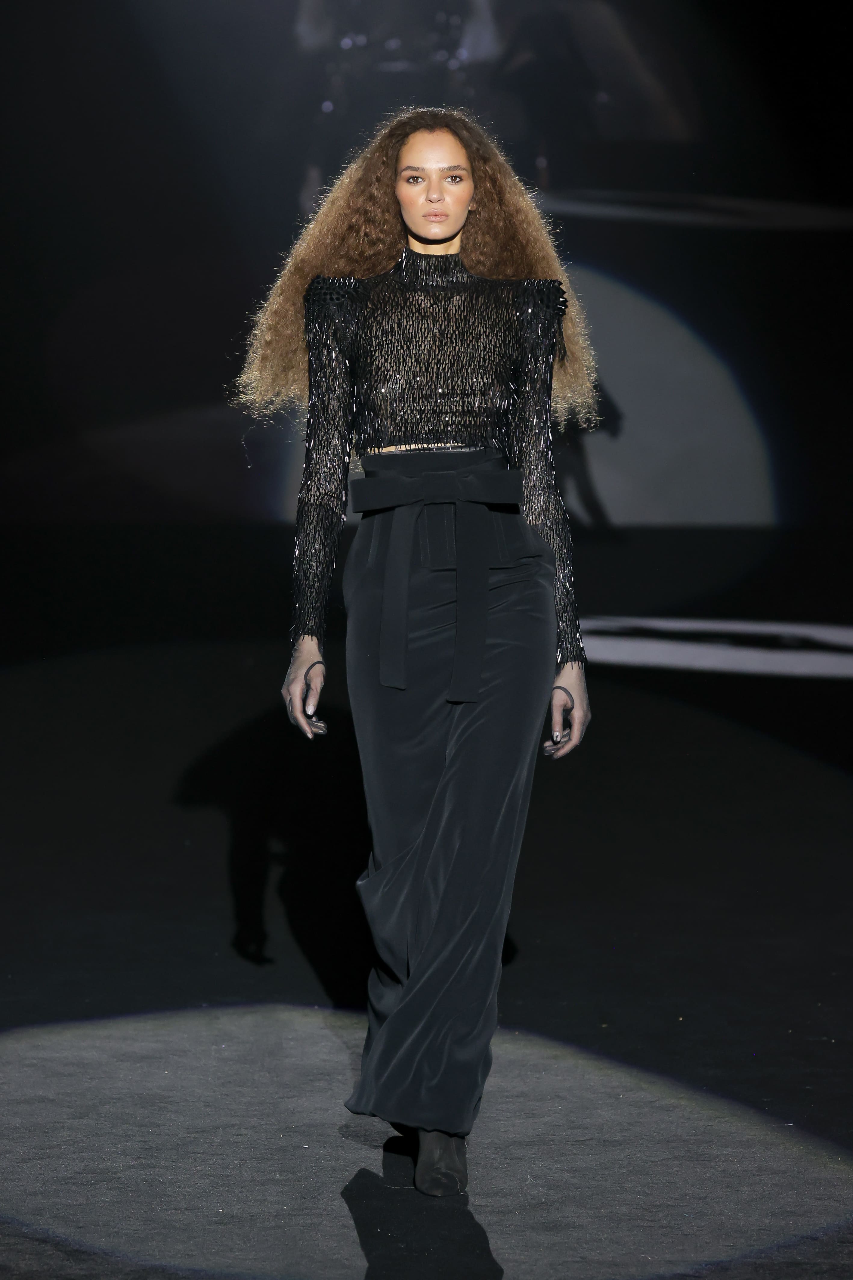 The model walks the runway in an elegant black outfit with a textured top and a long skirt.