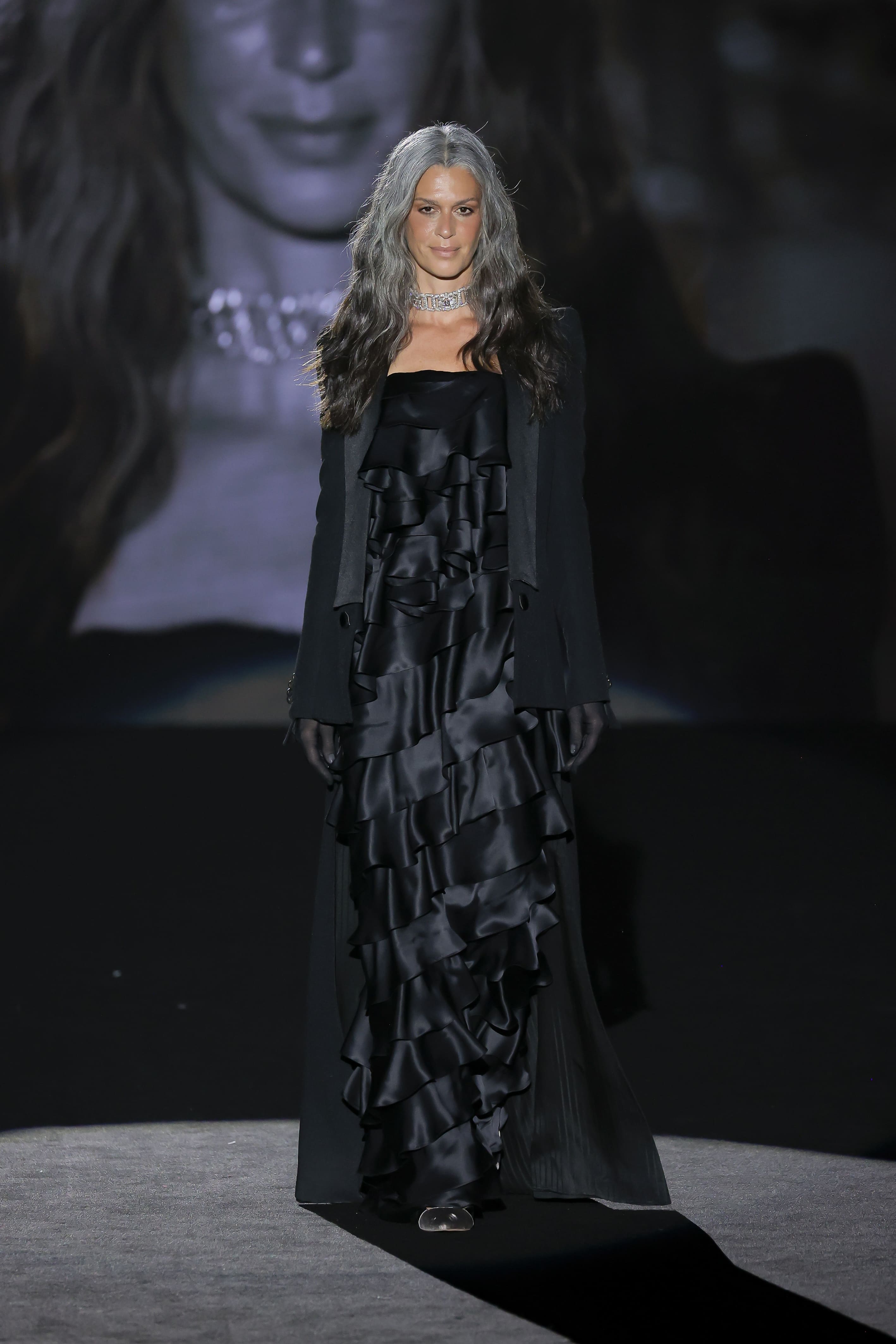 The model walks the catwalk in a long black dress with ruffles and gloves.