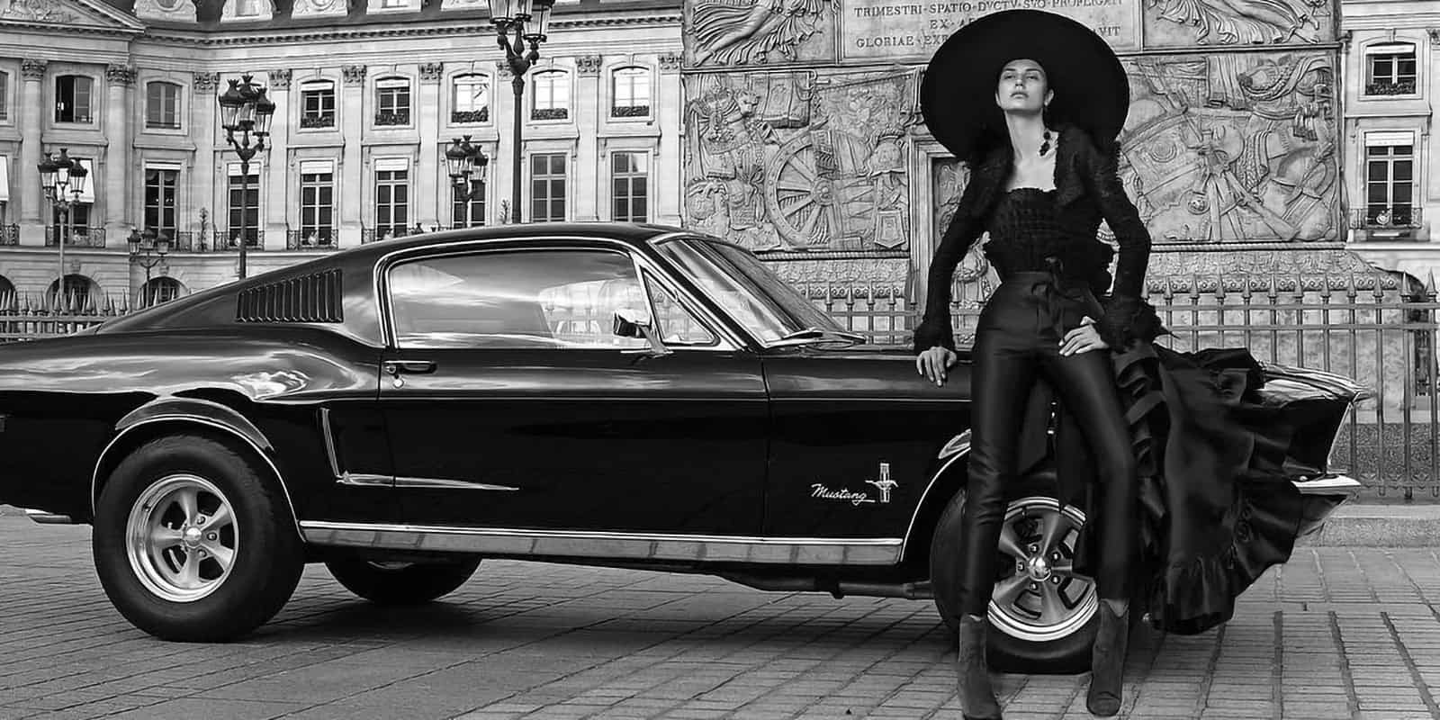 Modelo de Malne posa junto a un coche Mustang negro con sombrero ancho y look negro dramático en blanco y negro.