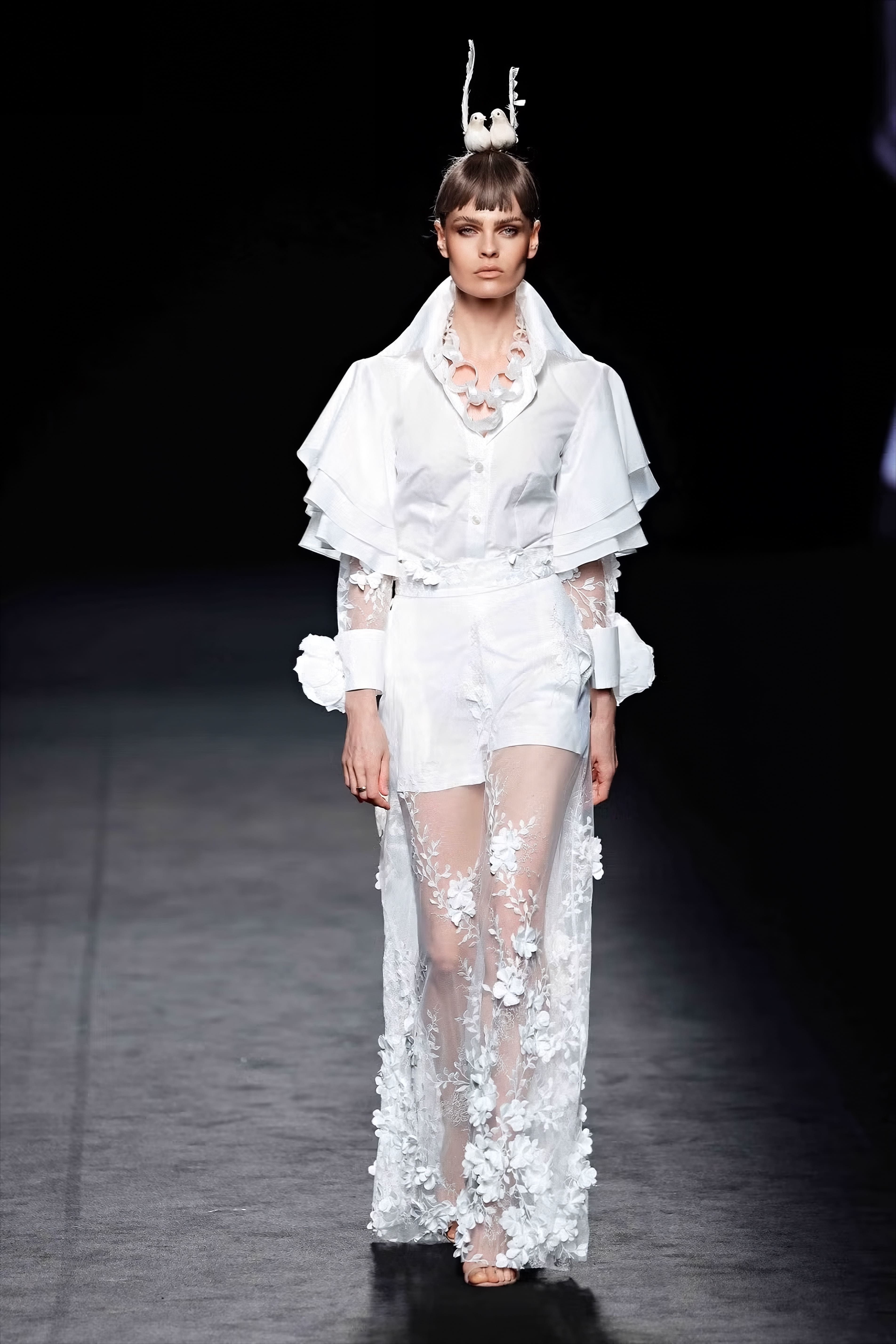 Modelo en la pasarela con conjunto blanco de Malne formado por camisa de mangas en capas, pantalón corto y sobrefalda larga semitransparente con bordados florales en relieve.