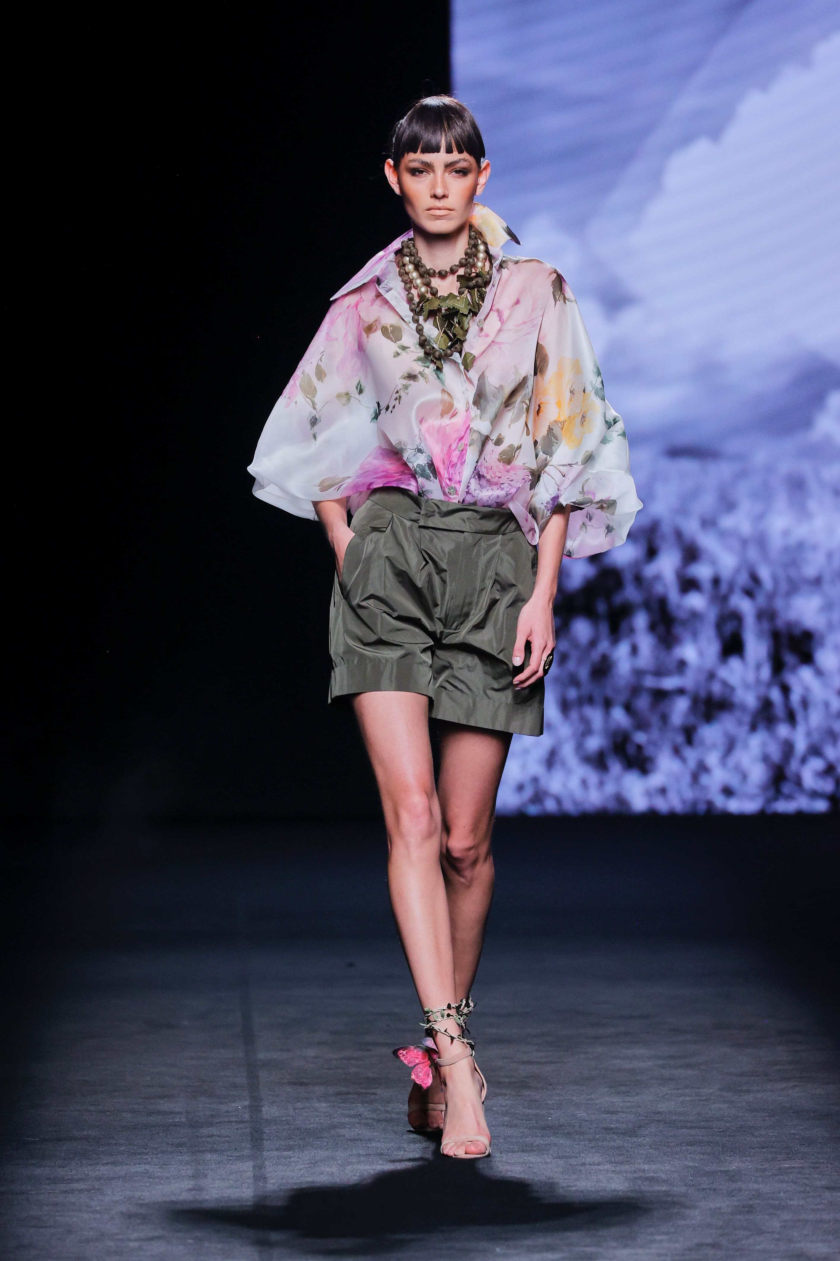Modelo desfila en la pasarela con blusa vaporosa estampada y falda corta verde.