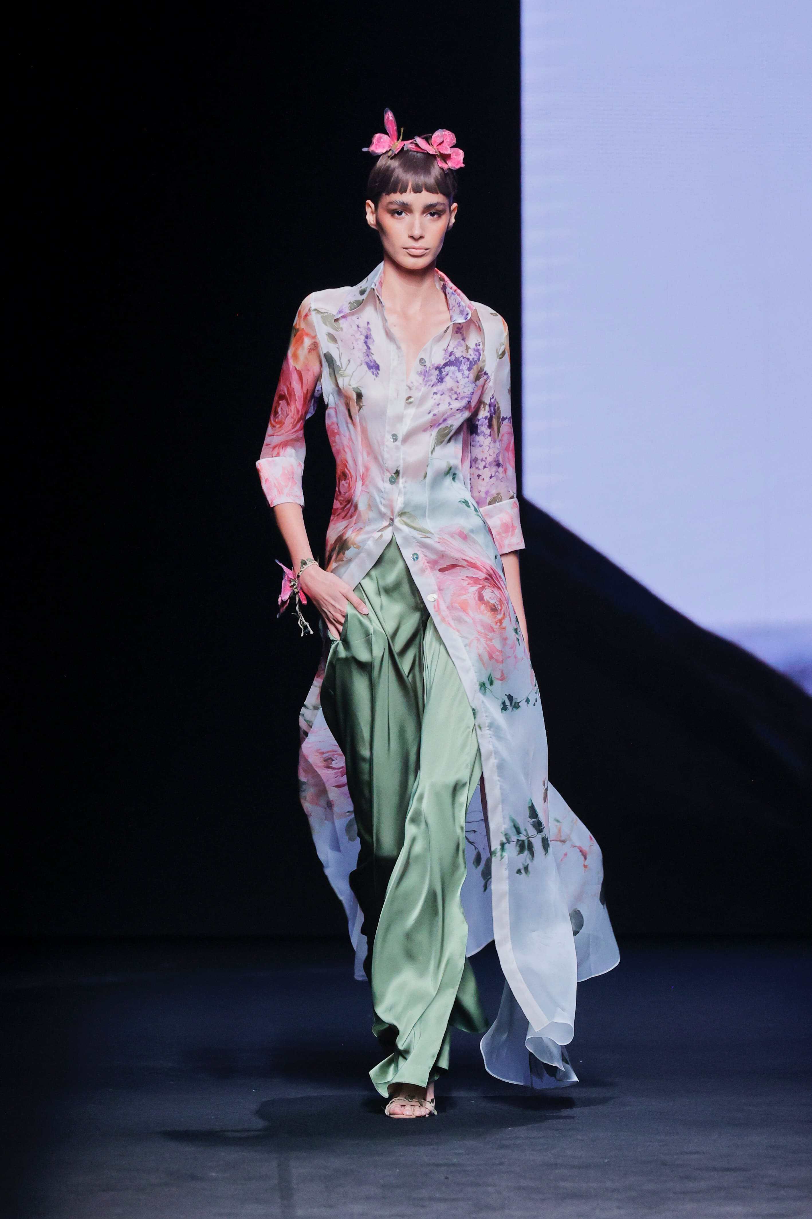 Modelo en la pasarela con conjunto de Malne formado por camisa larga semitransparente con estampado floral en tonos rosa y lila, combinada con pantalón fluido verde satinado.