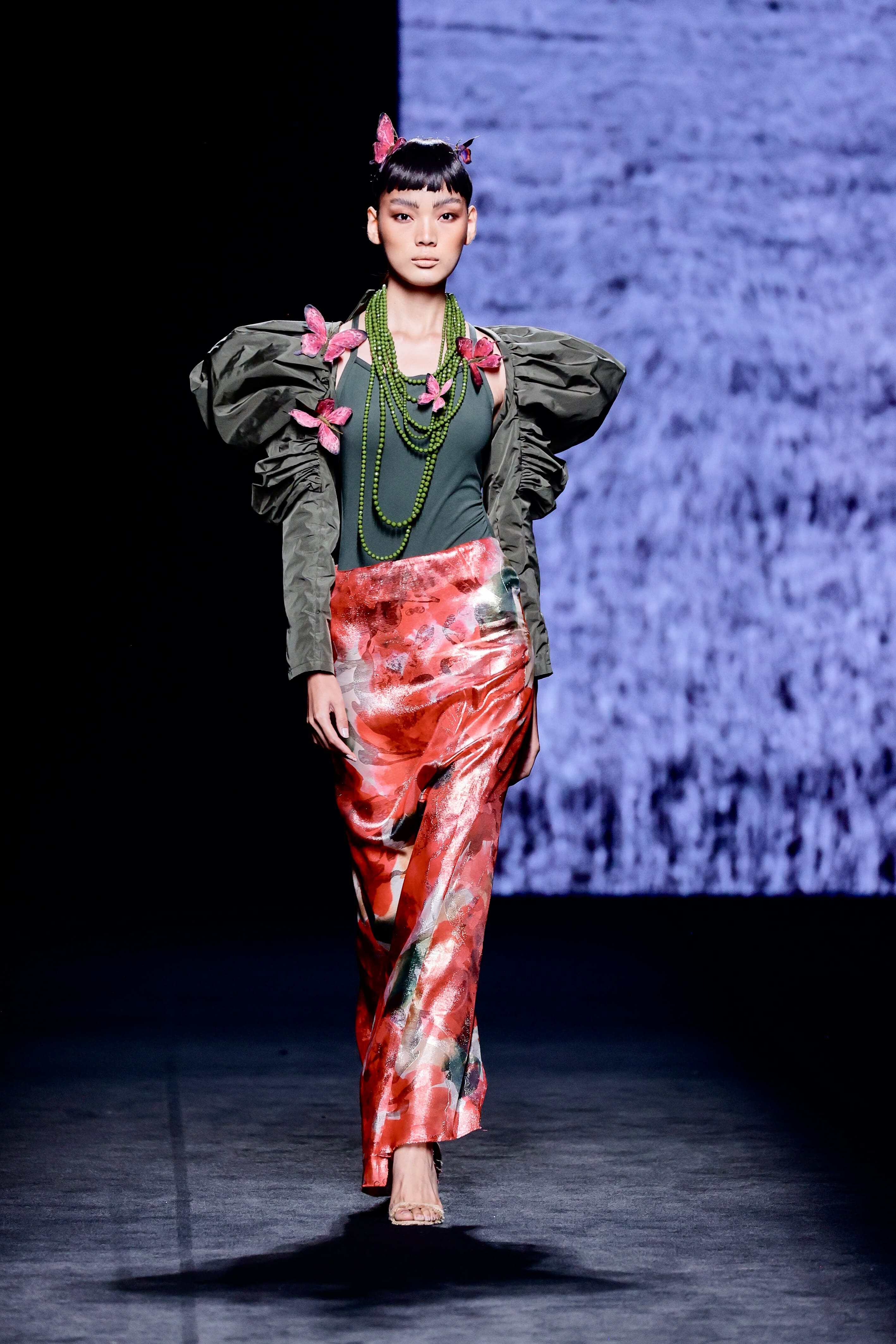 Modelo desfila en la pasarela con chaqueta verde de hombros marcados y falda larga satinada estampada.