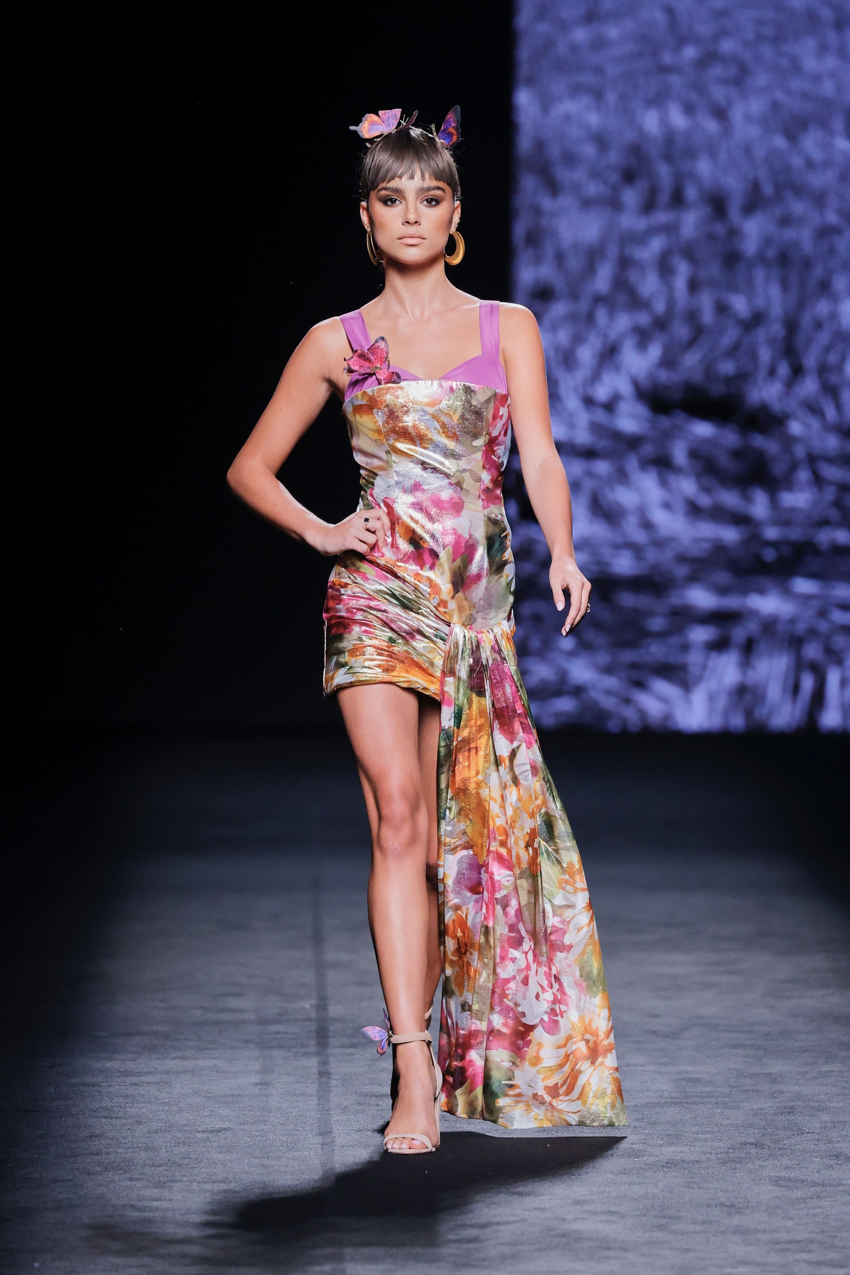 Modelo en la pasarela con vestido corto estampado de flores de Malne en tonos rosa, naranja y dorado, con tirantes anchos en color lila y drapeado lateral que cae en un panel largo.
