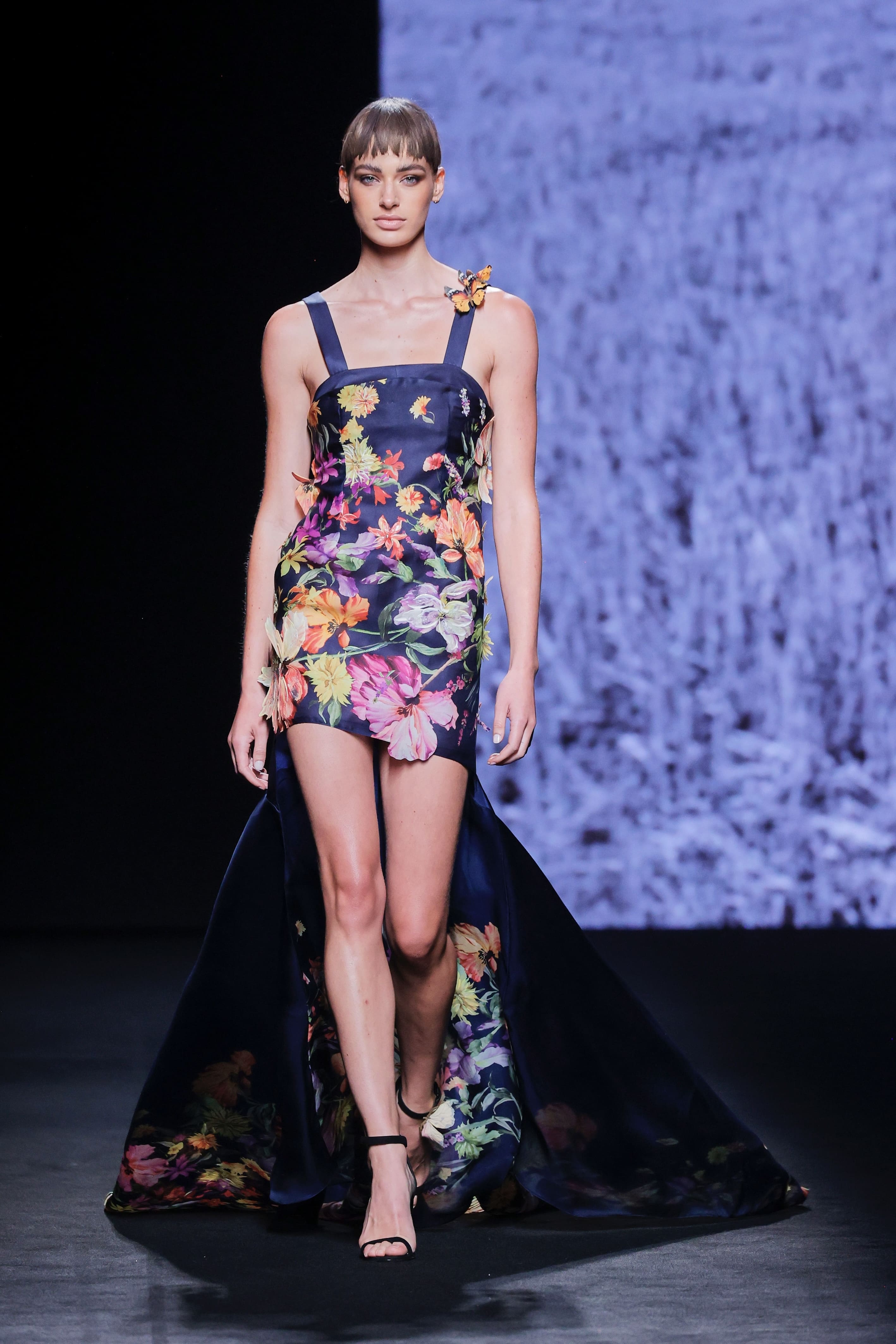 Modelo en la pasarela con vestido corto azul marino de Malne estampado con flores en tonos naranja, rosa y lila, con tirantes anchos y una pequeña mariposa en un hombro.