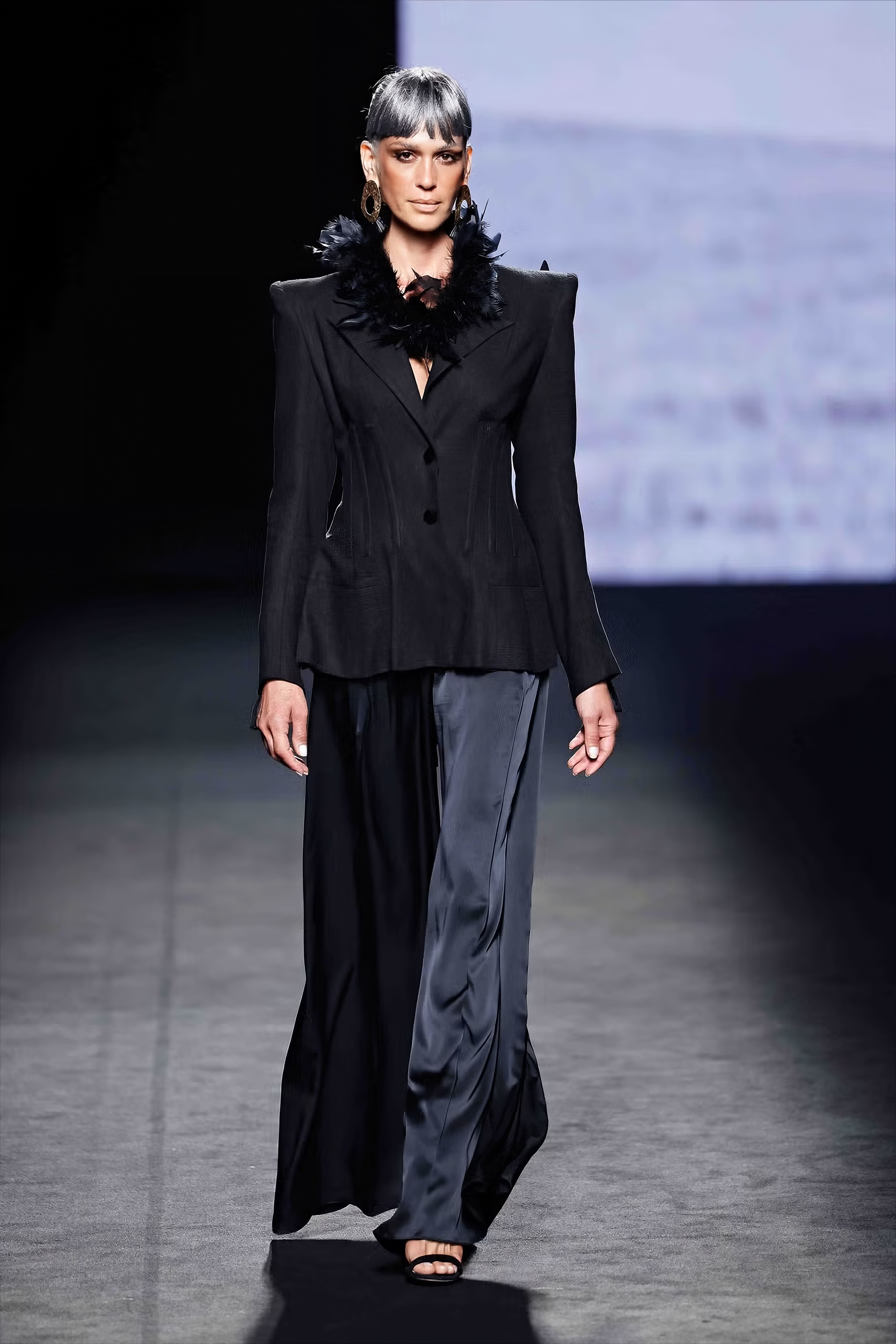 Modelo en la pasarela con conjunto negro de Malne compuesto por americana estructurada con hombros marcados y pantalón fluido de acabado satinado.