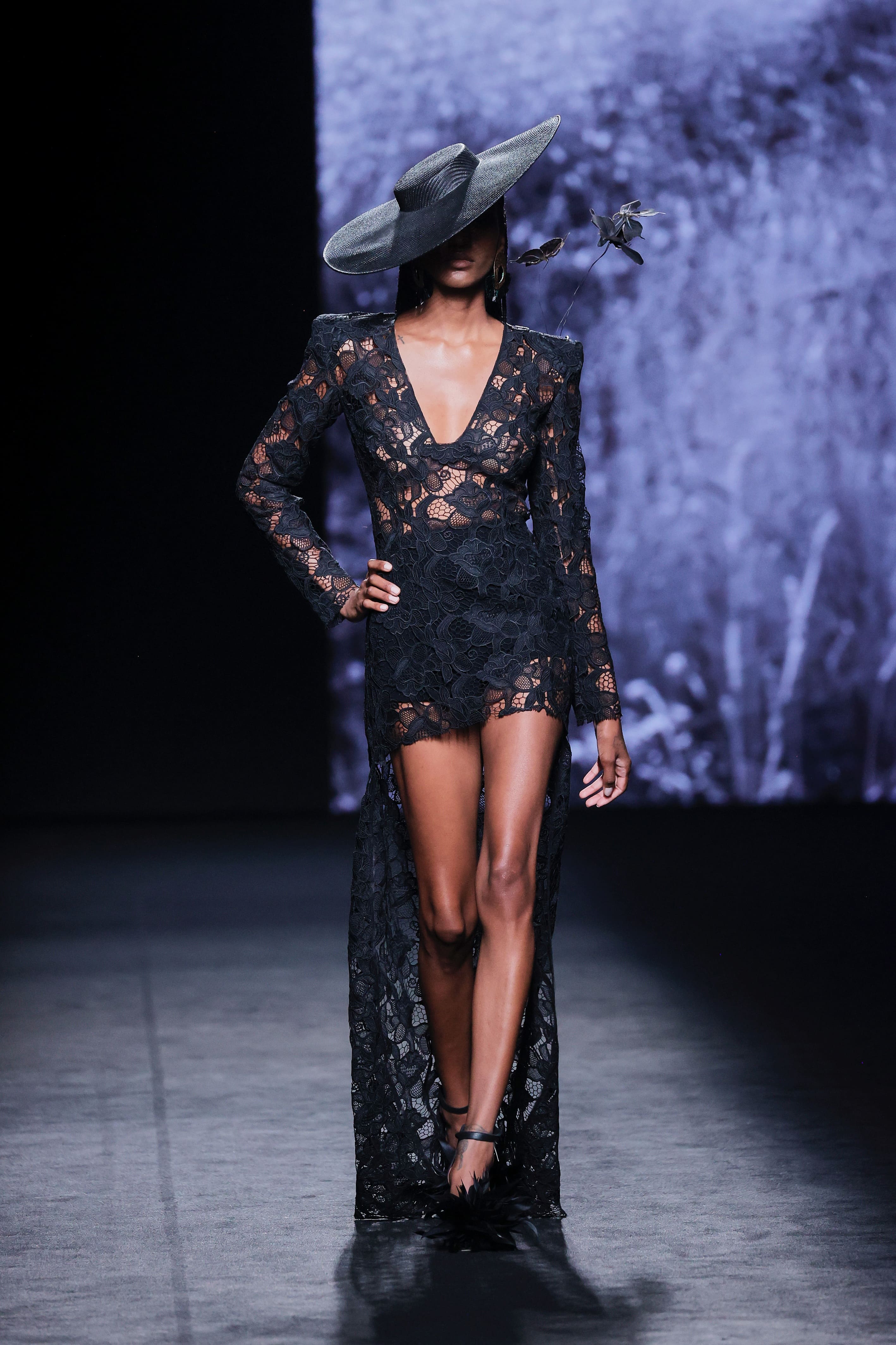Modelo en la pasarela con mono corto negro de encaje de Malne, con escote profundo, hombros estructurados y sobrefalda larga semitransparente.