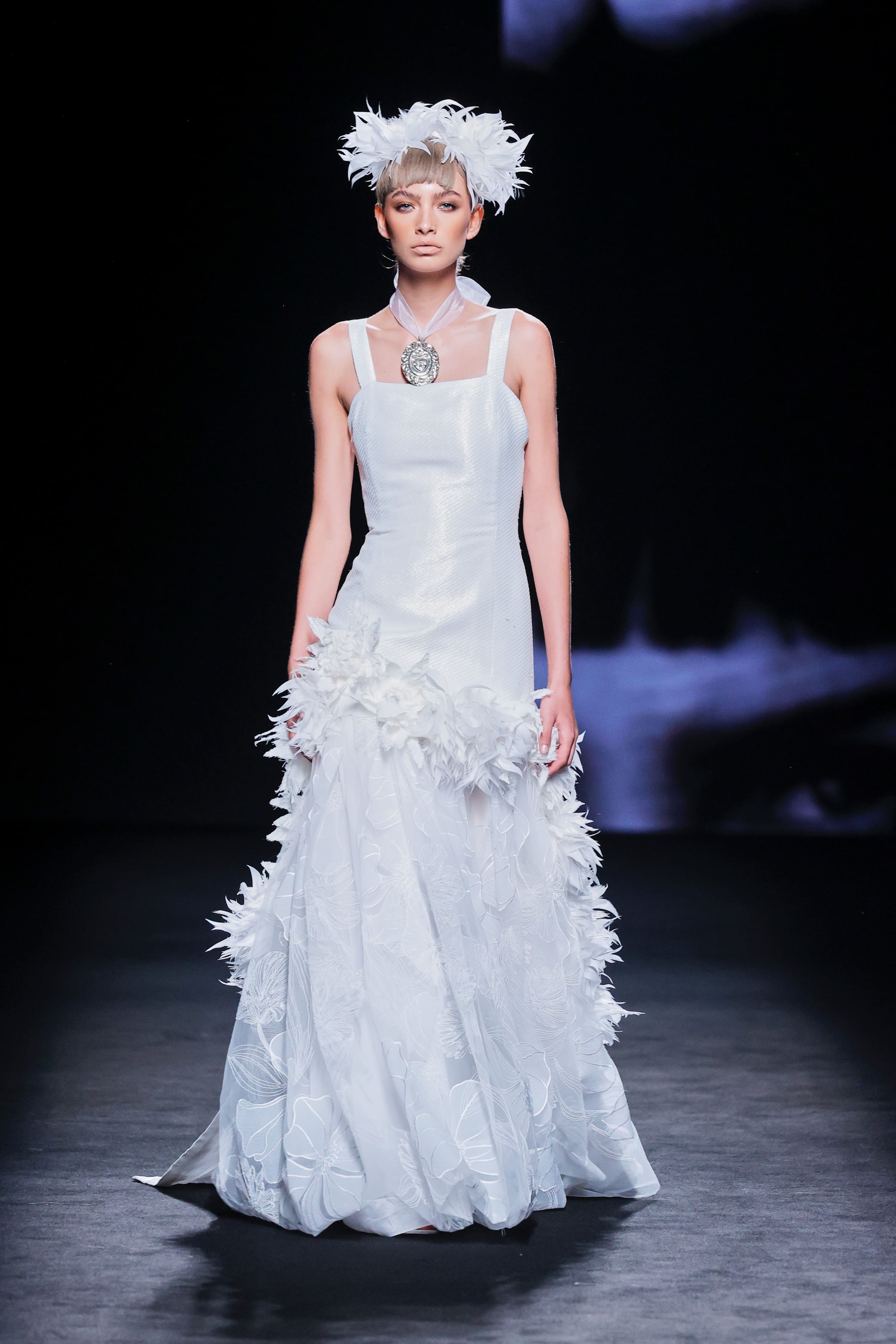 Modelo desfila en la pasarela con un vestido largo blanco con detalles de plumas y tocado a juego.