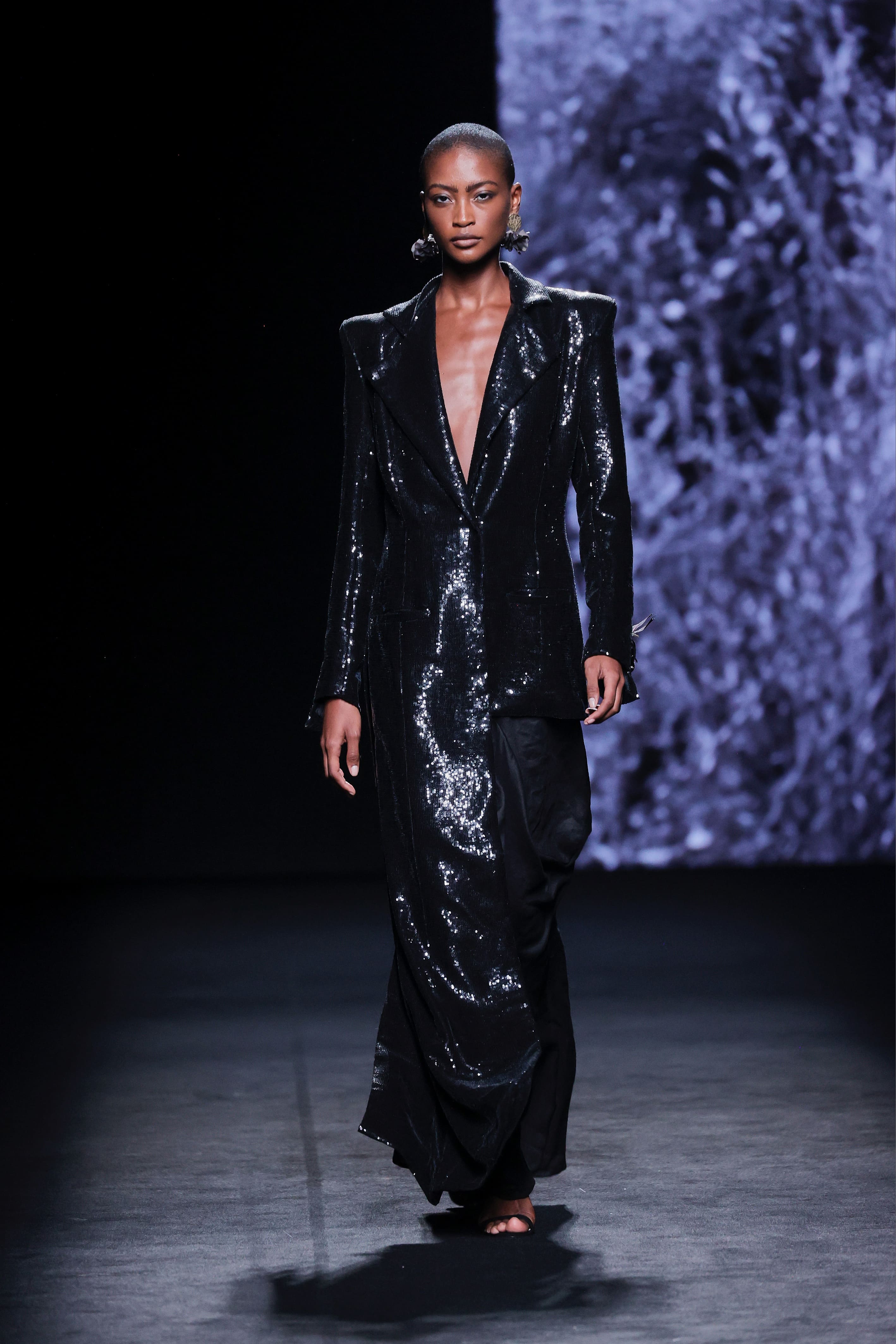 Modelo en la pasarela con vestido negro de Malne de acabado brillante y corte blazer con hombros marcados y escote profundo.