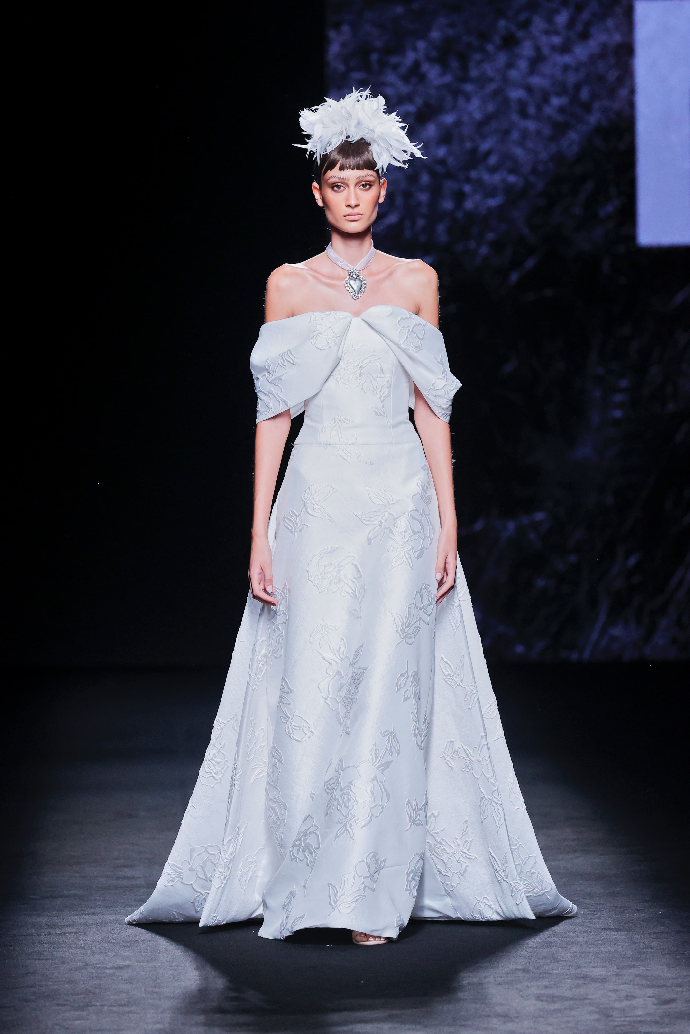 Modelo desfila en la pasarela con un vestido largo blanco de hombros descubiertos y tocado de plumas.