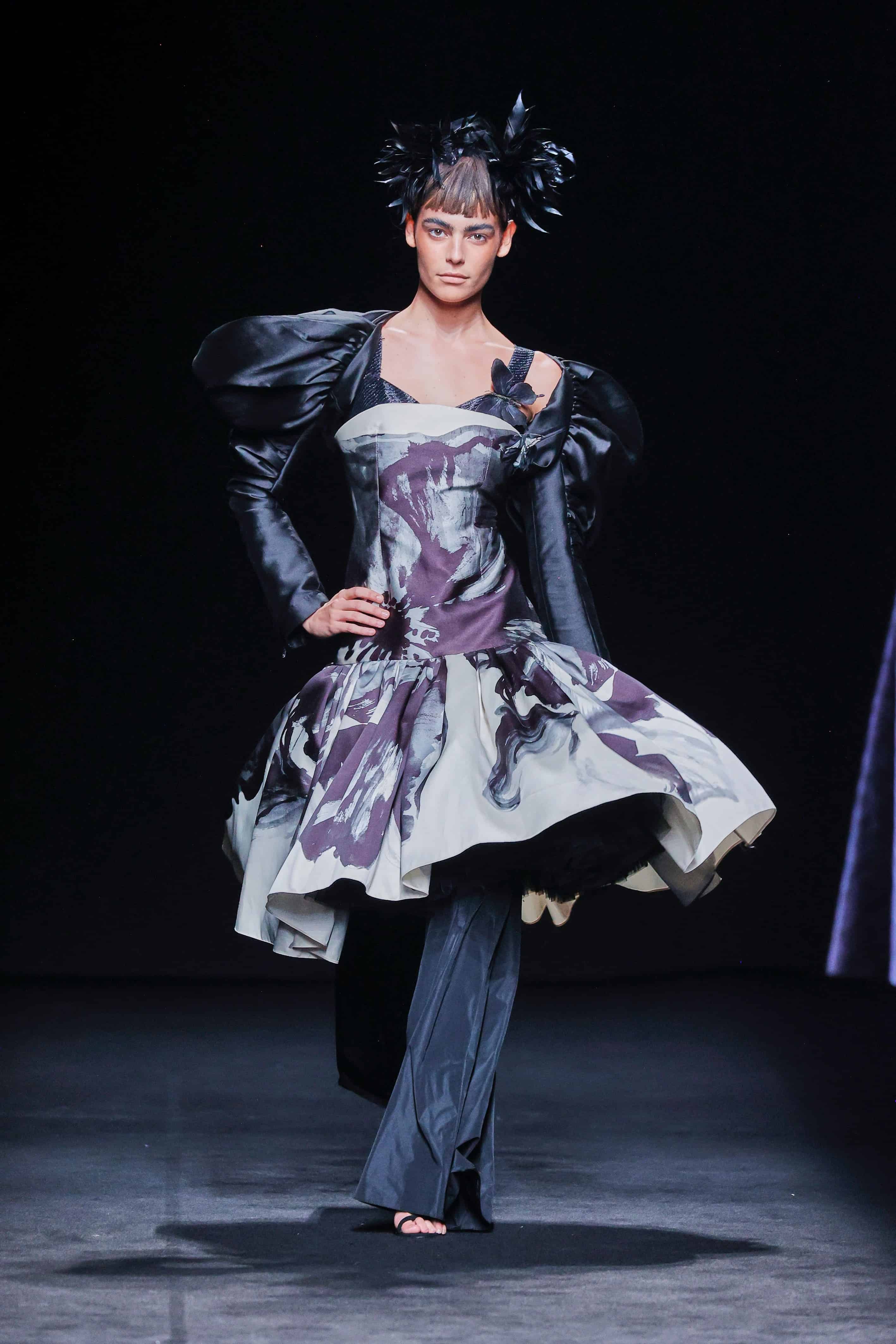 Modelo en la pasarela con vestido estructurado de Malne en tonos blanco, negro y morado, con falda amplia de vuelo y mangas negras abullonadas.