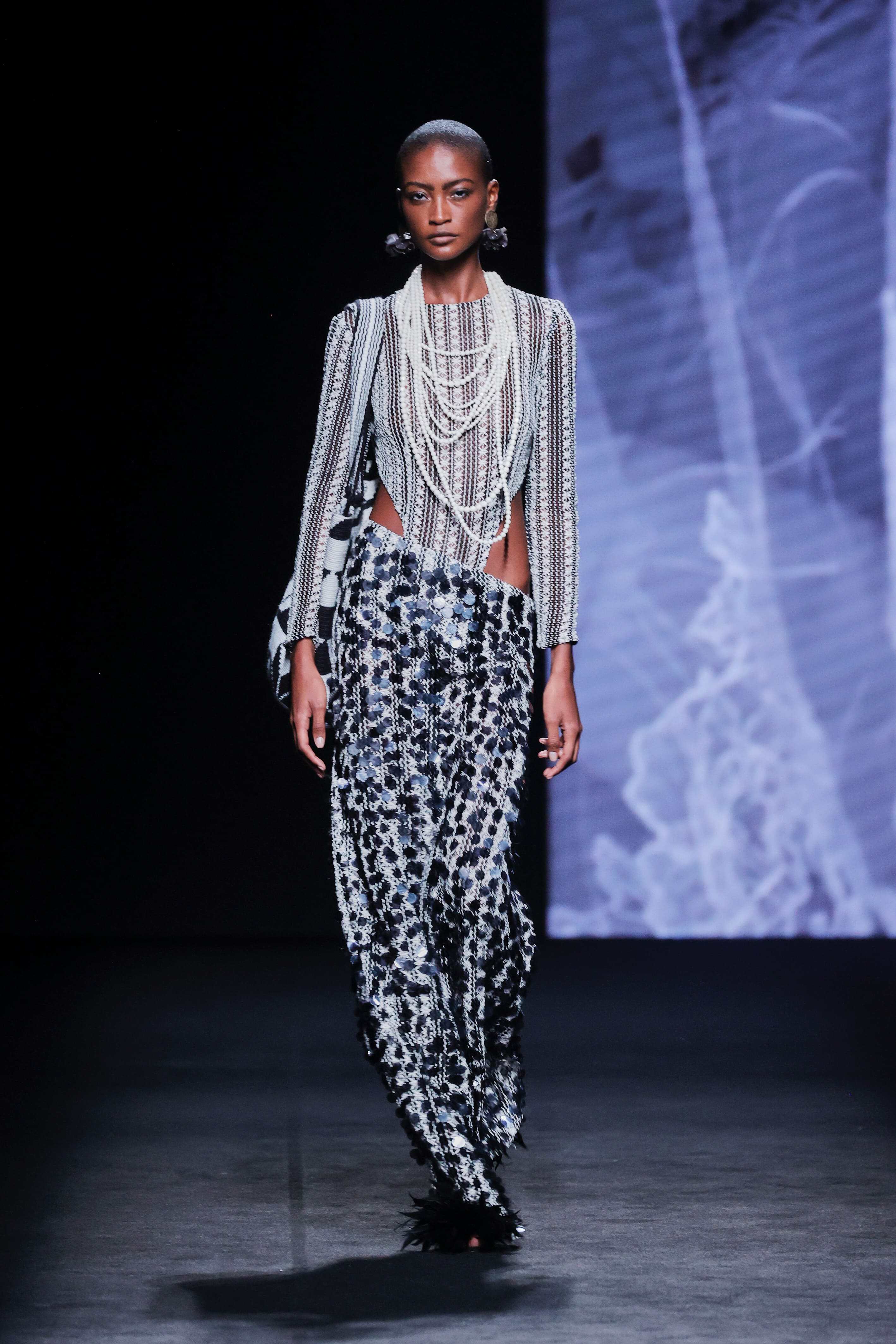 Modelo en la pasarela con conjunto de Malne compuesto por top de mangas largas cubierto de cadenas plateadas y falda larga de lentejuelas en negro y plata.