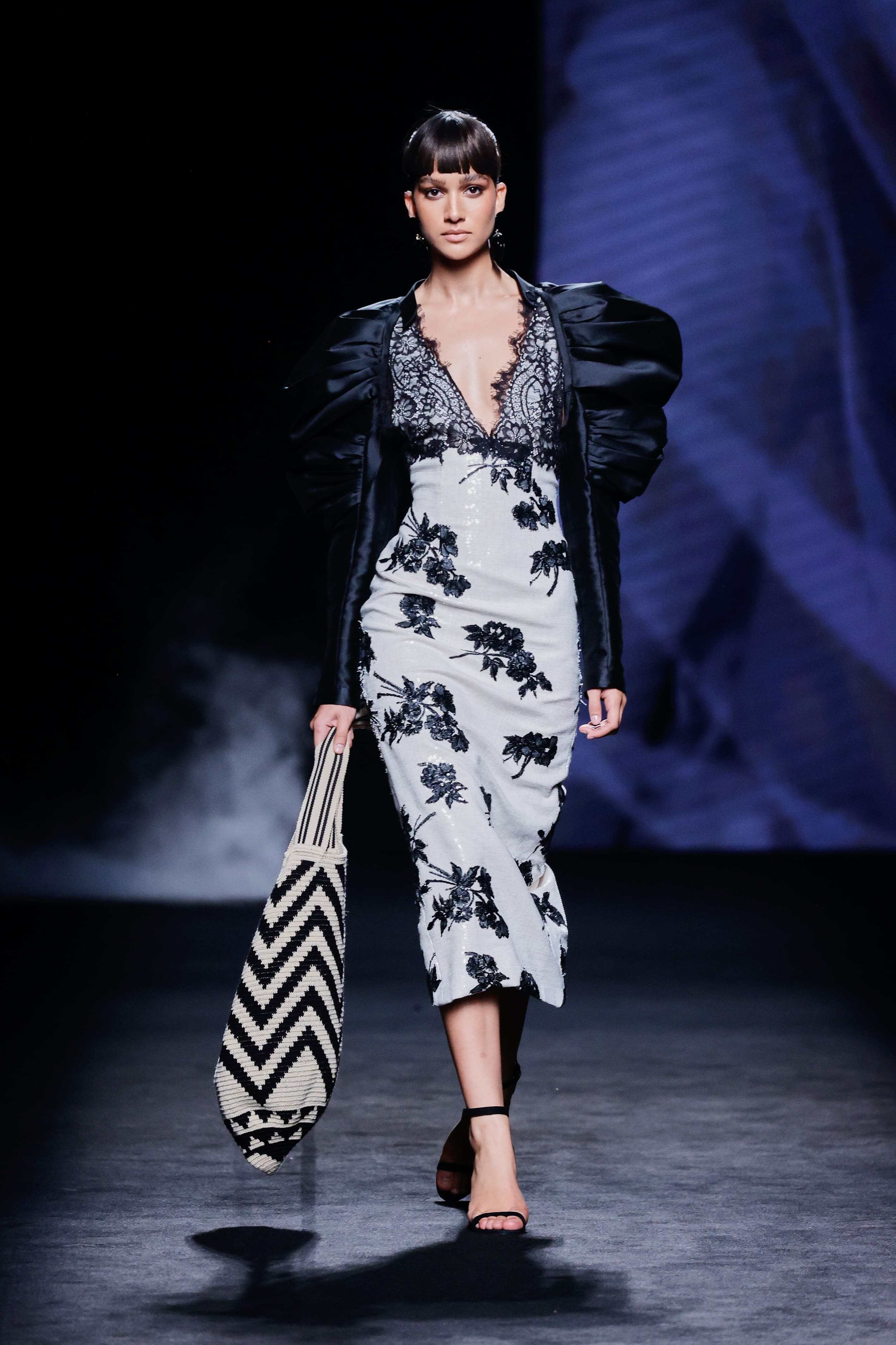 Modelo en la pasarela con vestido midi blanco de Malne decorado con bordados florales negros y escote profundo de encaje negro.