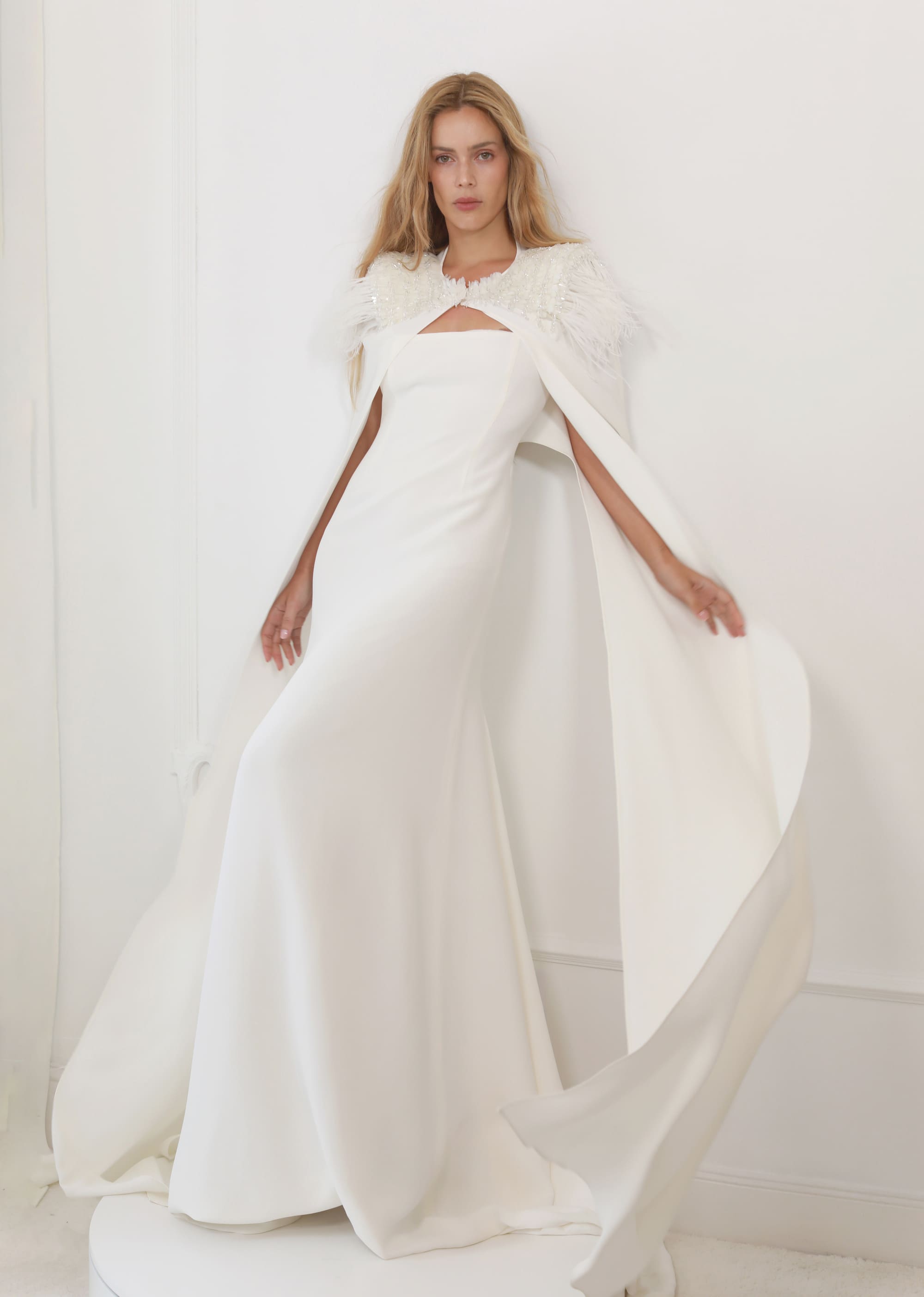 Model in a fitted white dress with a straight neckline, accompanied by a cape with feathers and shiny appliqués on the shoulders.