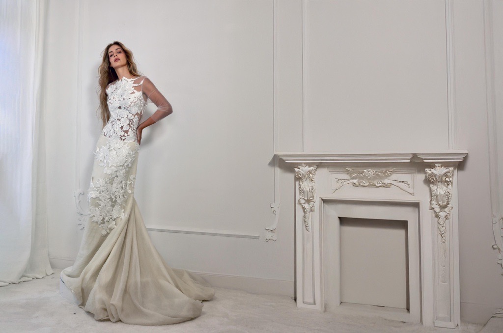Modelo con vestido de novia ajustado en tono marfil, con aplicaciones florales en relieve, transparencias en mangas y escote, y falda con cola de sirena