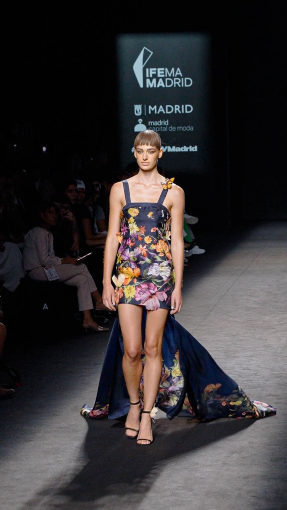 Modelo de Malne en la pasarela de IFEMA con minivestido floral y cola larga sobre fondo oscuro.