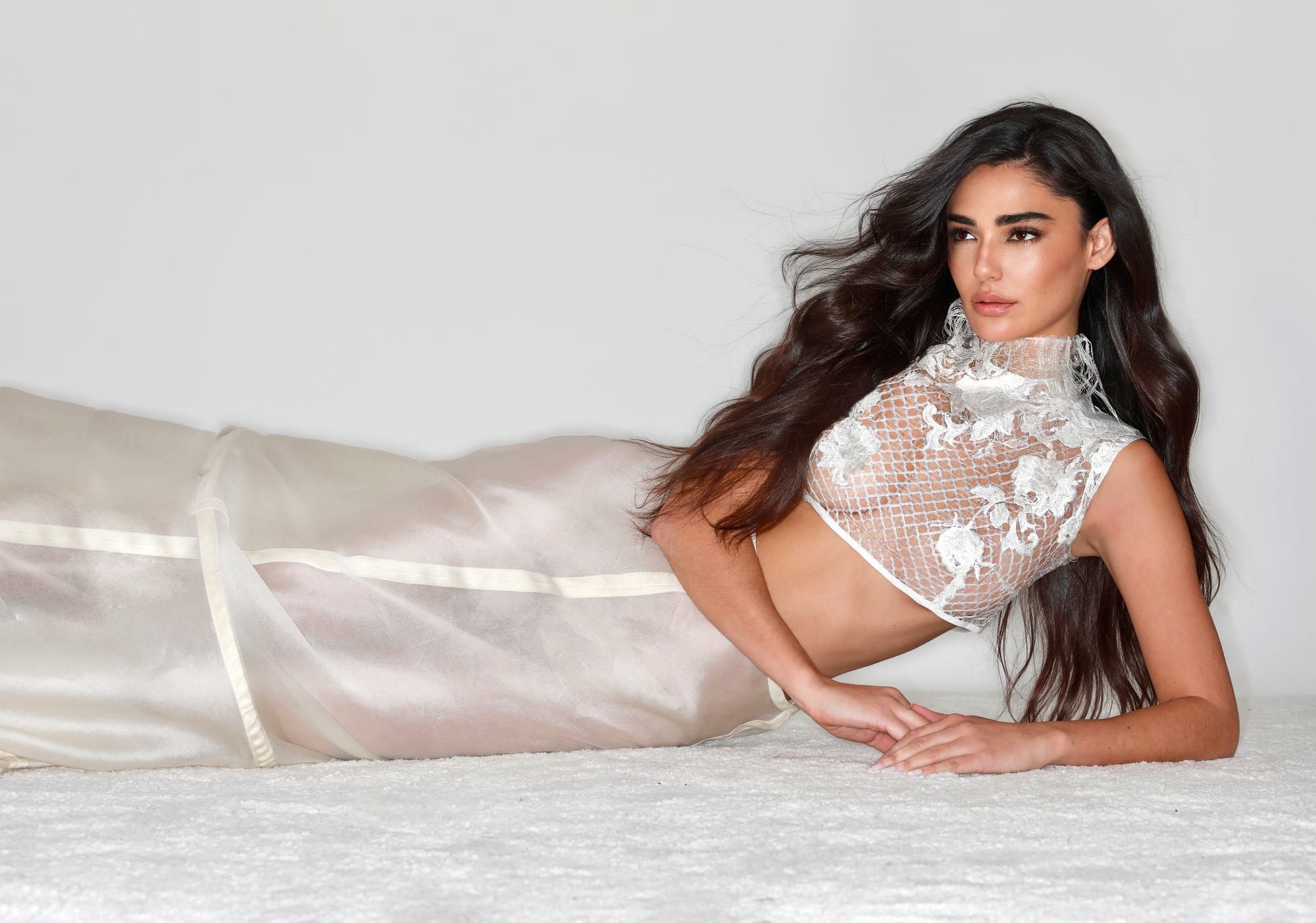 A woman lies on a light surface wearing a long nude-coloured dress in a semi-transparent fabric. The design features embossed floral embroidery at the top and a fitted silhouette that highlights the elegance and delicacy of the ensemble.