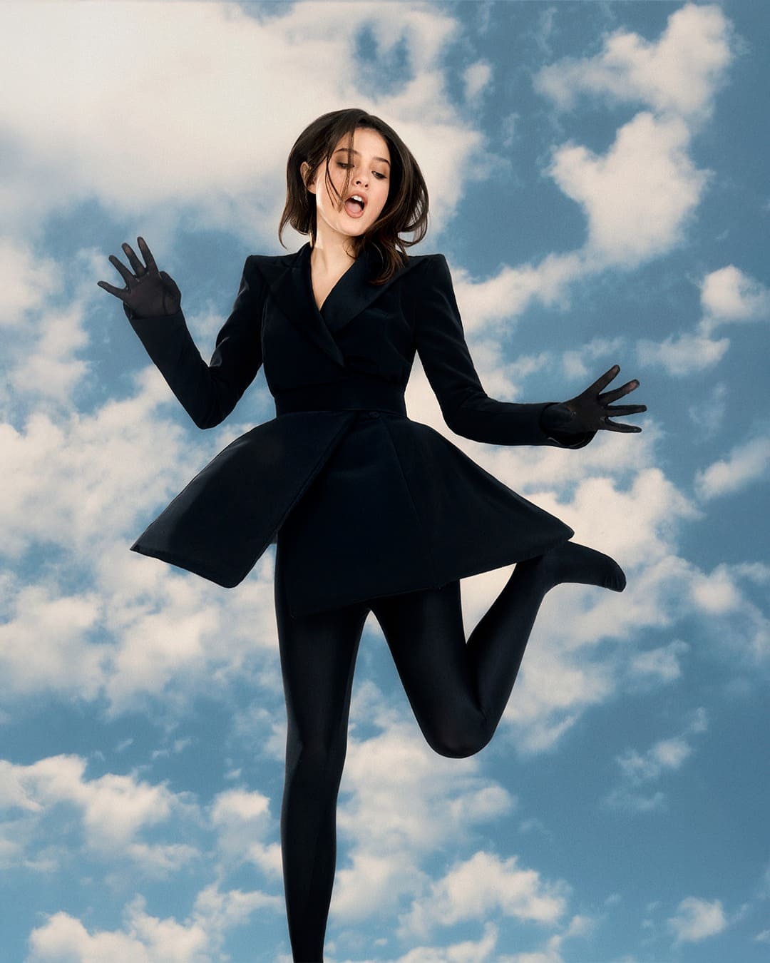 Woman jumps in the air in front of a blue sky with white clouds while wearing a structured black outfit with short flared skirt, gloves and tight stockings.