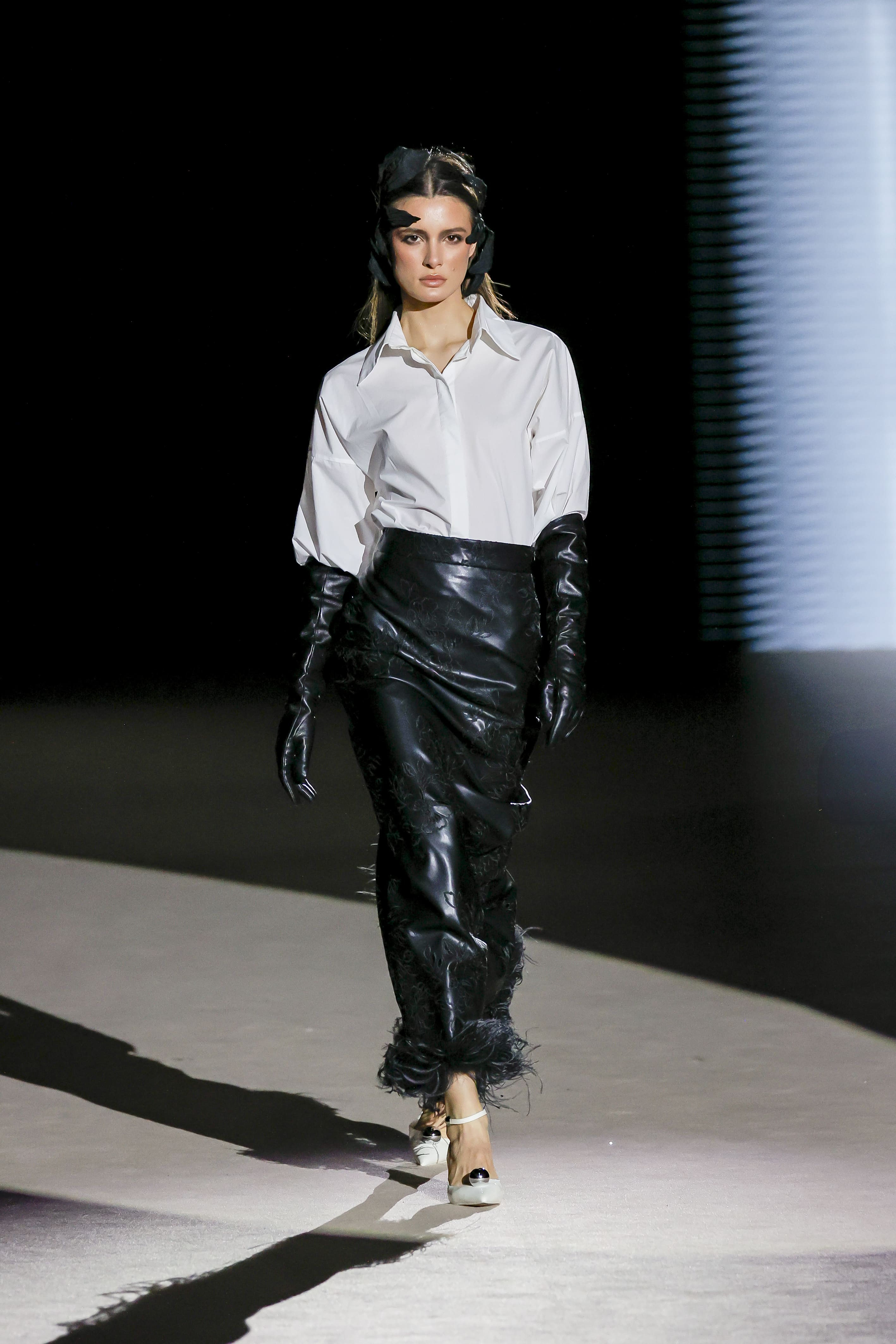 Modelo desfilando con un diseño de Malne en la Mercedes-Benz Fashion Week Madrid en marzo de 2026 con camisa blanca y falda negra de acabado brillante con guantes largos.