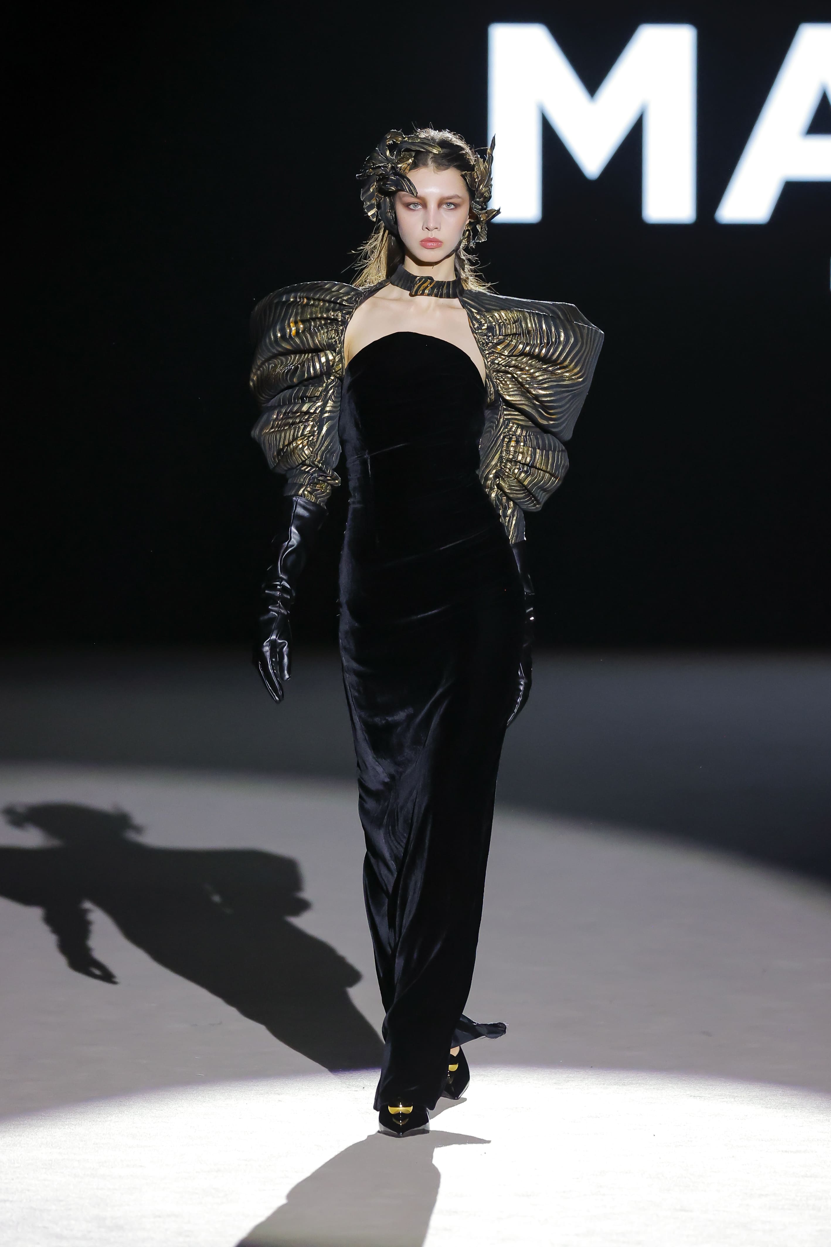 Modelo en pasarela con vestido negro largo de Malne, mangas abullonadas doradas y guantes negros.