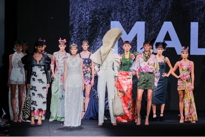 Modelos en pasarela presentan la colección de Malne con vestidos florales, lentejuelas y accesorios llamativos sobre un escenario oscuro.